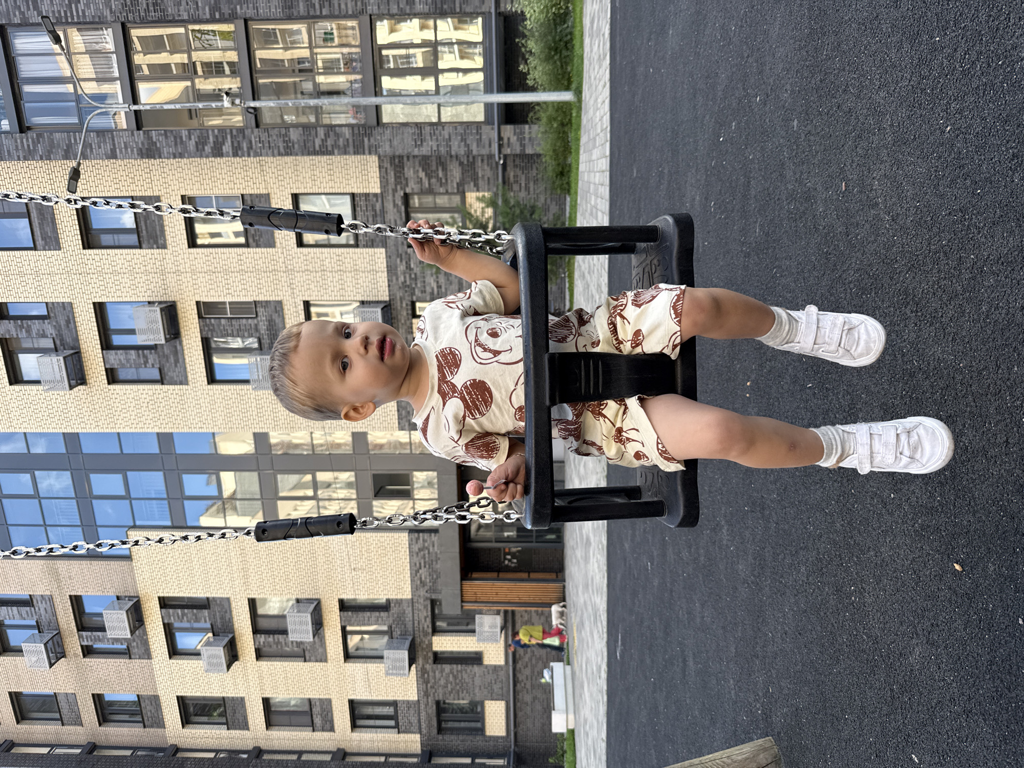 A young Caucasian child with light brown hair is seated on a black swing in an urban playground. The child is wearing a light-colored t-shirt and shorts with a brown patterned print and white sneakers. The child is holding a small pink lollipop in their right hand. The swing set is in the foreground, with a dark gray rubberized playground surface beneath it. In the background, a modern apartment building with a beige brick facade and dark gray window frames stands prominently. To the right of the building, a street lamp is visible. Further to the left, in the mid-ground, two adults and a dog are walking away from the camera. One adult is wearing a yellow top and red pants, while the other is wearing a dark top and pants. The setting appears to be the city of Rusanovo, Russia, during daylight hours.