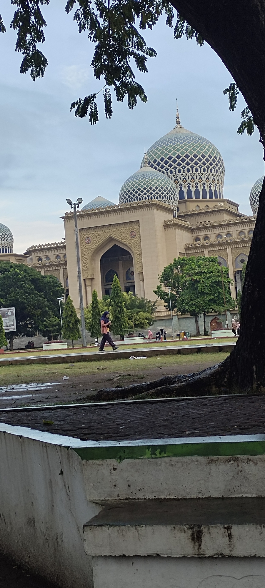The image depicts the Baiturrahman Grand Mosque in Lhokseumawe, Indonesia, under an overcast sky. The mosque, a prominent landmark, features multiple domes, the largest of which is adorned with intricate green and white geometric patterns. The building itself is light-colored stone with arched entrances and decorative carvings. In the foreground, a young woman wearing a hijab and a maroon top with dark pants walks across a grassy area, holding a mobile phone. Behind her, a few other individuals are seated or standing on a paved walkway. Trees, some with lush green foliage, are scattered around the mosque and the surrounding grounds. A large tree trunk is visible in the right foreground, partially obscuring the view of the mosque. The ground in the immediate foreground appears wet and somewhat muddy, suggesting recent rain. The overall scene is tranquil, with the mosque serving as the dominant architectural element.