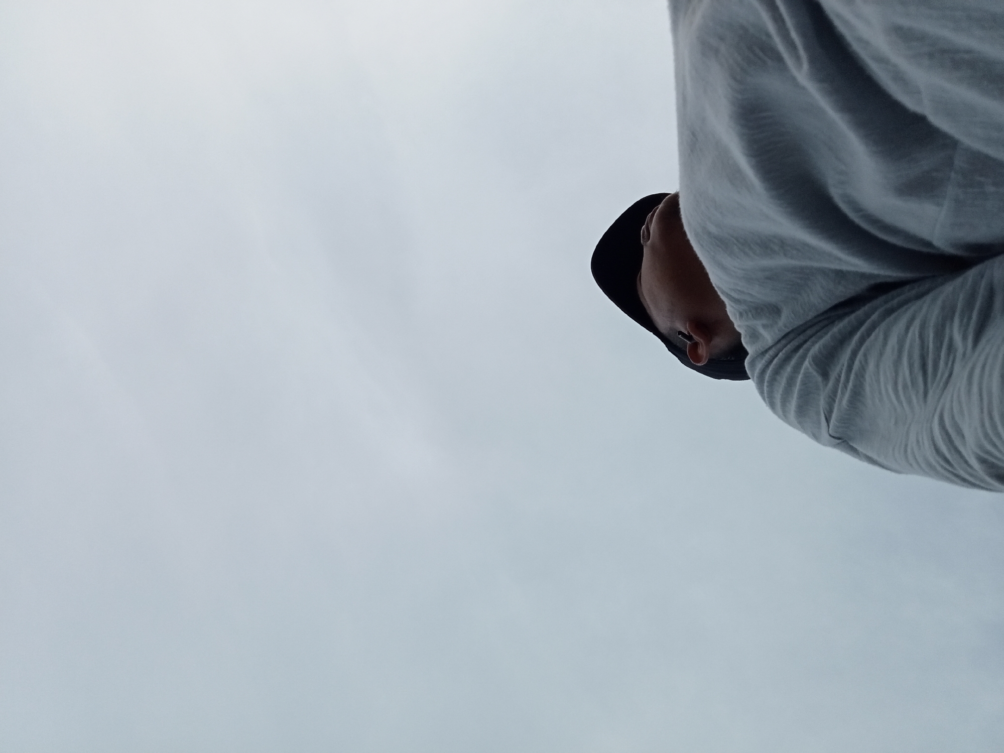 The image, taken from a low angle, shows a person wearing a grey long-sleeved shirt and a black hat. The person's face is visible, looking downwards, and part of their neck and shoulder are also visible. The background is a uniformly overcast sky, suggesting an outdoor setting, possibly a public space in Kualu, Indonesia, though no specific landmarks or urban features are identifiable. The lighting is diffused, indicating it is daytime but overcast. There are no other people or objects in the frame.