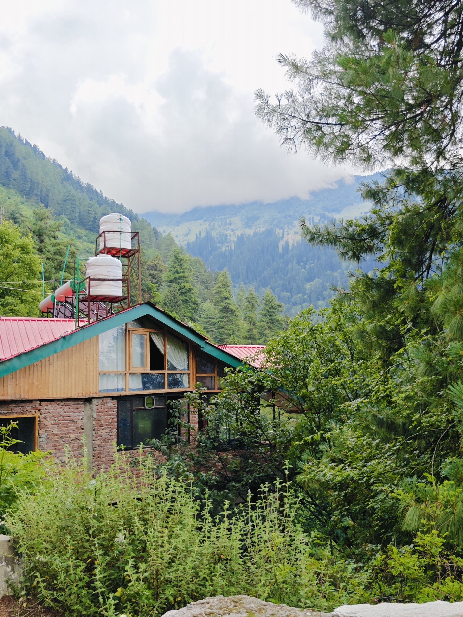 The image depicts a multi-story building constructed with brick and wood in Prini, India. The building has a red tin roof with green trim. On the roof, there are two large white water tanks supported by a metal structure, and a red cylindrical tank. The building features several windows, some with wooden frames and curtains visible. Surrounding the building is lush green vegetation, including trees, bushes, and smaller plants. In the background, a densely forested mountain range rises under a cloudy sky. The scene appears to be a residential or guesthouse structure situated in a mountainous, verdant environment.