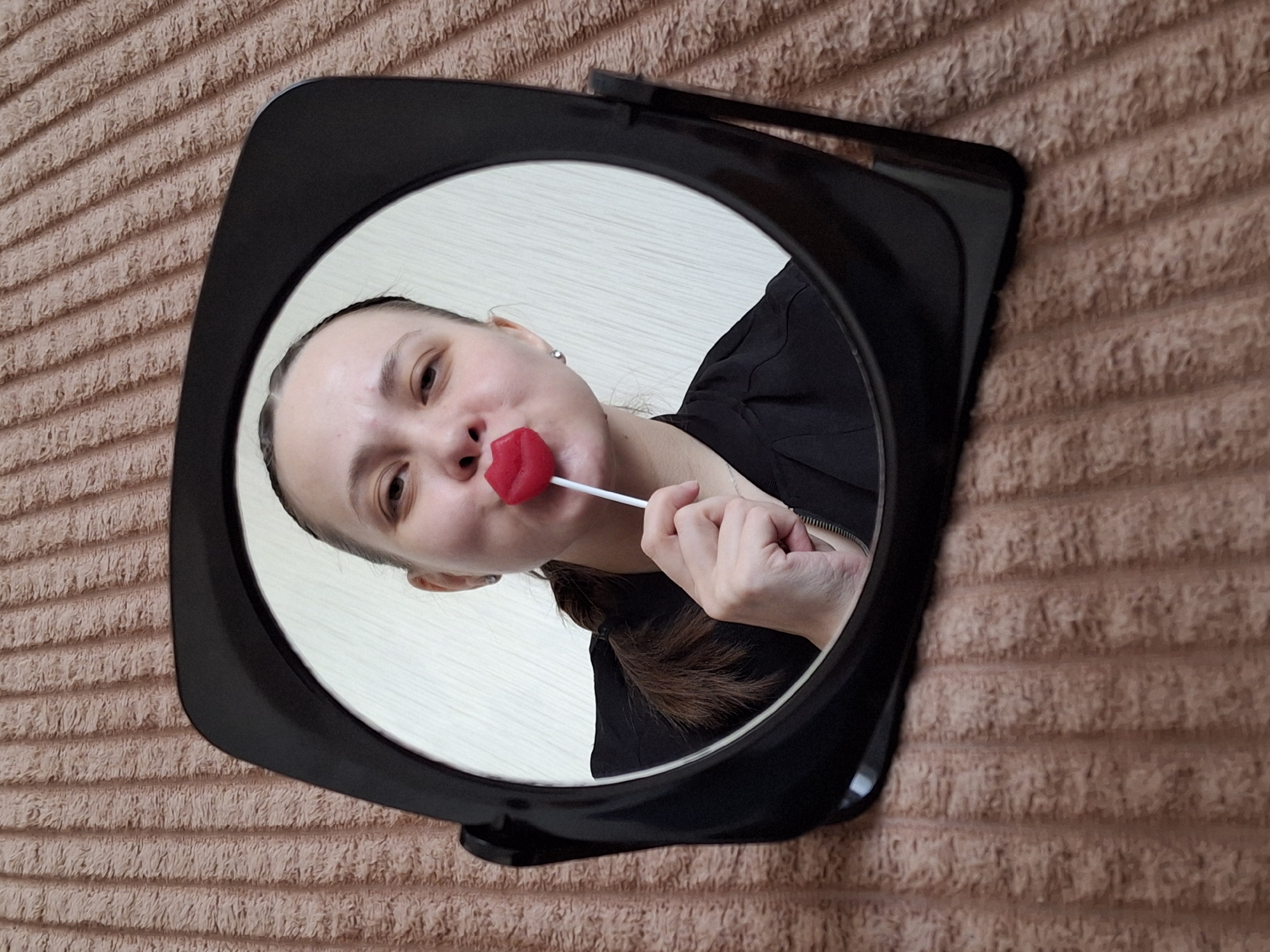 A woman is visible in the reflection of a black, round-shaped mirror. She is holding a red, lip-shaped lollipop to her mouth with her right hand. The woman has her brown hair pulled back in a ponytail and is wearing a black shirt. The mirror is placed on a brown, horizontally-lined fabric surface. The image was taken in Chaikovsky, Russia.