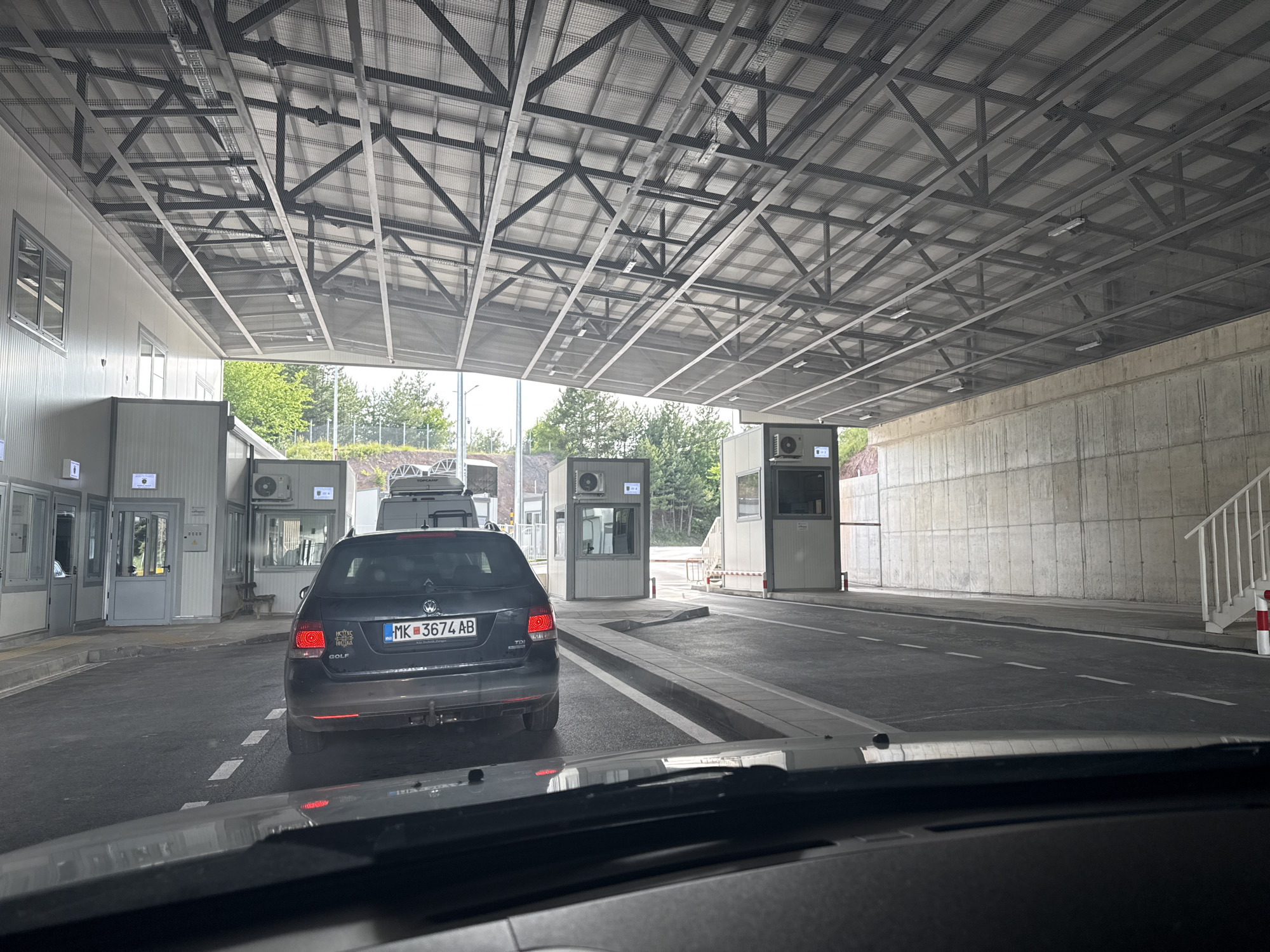 The image depicts a border crossing station in the city of Klisura, Bulgaria, viewed from the perspective of a vehicle stopped in traffic. A dark-colored Volkswagen Golf with license plate MK 3674 AB is in the foreground, stopped in the right lane of a paved road. The vehicle has its brake lights illuminated. Ahead of the Golf, a series of white booths are visible, presumably for border control. These booths are situated under a large, metal-framed canopy structure with a mesh ceiling. To the right of the booths, a concrete wall extends into the distance, with a white staircase featuring red and white striped posts. In the background, trees and foliage are visible, suggesting an outdoor environment. The overall scene appears to be a functional border crossing, with infrastructure designed to manage vehicle and pedestrian traffic. There are no identifiable individuals in the immediate foreground, but the presence of multiple vehicles and the border control booths implies human activity at the location. The lighting suggests it is daytime, with natural light filtering through the canopy and illuminating the scene.