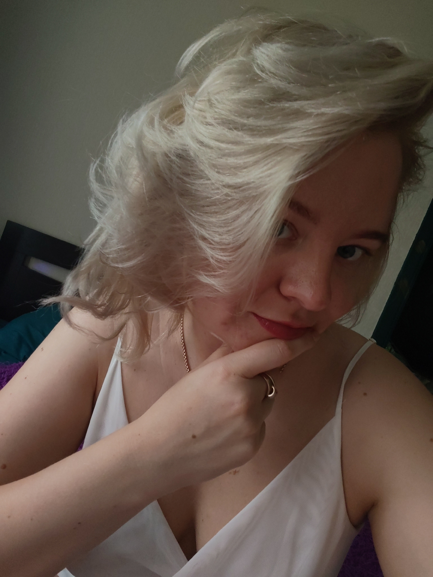 A young woman with light blonde hair is posing for a selfie indoors. She is wearing a white sleeveless top and a gold necklace. Her left hand is raised, with her index finger touching her chin. She is also wearing a ring on her finger. The background includes a dark headboard, a blue pillow, and a purple blanket or sheet. There is nothing in the image to indicate that it was taken in Izhevsk, Russia.