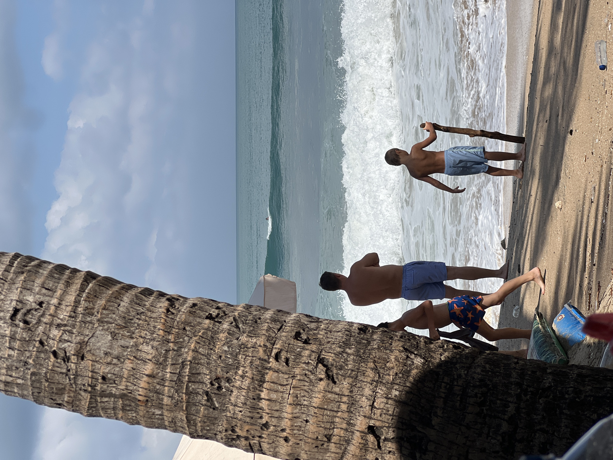An outdoor medium shot shows three people on a sandy beach in Kuta, Indonesia, with a large palm tree in the foreground obscuring part of the left side of the frame. The ocean with waves is in the background, and the sky is partly cloudy.
In the foreground, to the left and partially obscured by the palm tree, is a young boy wearing blue and orange patterned swim trunks. He is facing away from the camera and to the left, with his left arm raised as if he is reaching for the tree. Next to him, to his right, is a man in blue swim trunks, also facing away from the camera and slightly to the left. His arms are crossed. To the far right of the frame, a second young boy is standing on the sand, facing away from the camera and towards the ocean. He is wearing light blue swim trunks and holding a piece of driftwood vertically in his right hand.
The sand appears dry with some shadows cast from the palm tree. There are a couple of bags and a plastic water bottle on the sand. The ocean has white foamy waves crashing on the shore, and a small surfer is visible in the distance on a wave. A part of a beach umbrella is visible behind the man and to the right of the palm tree.