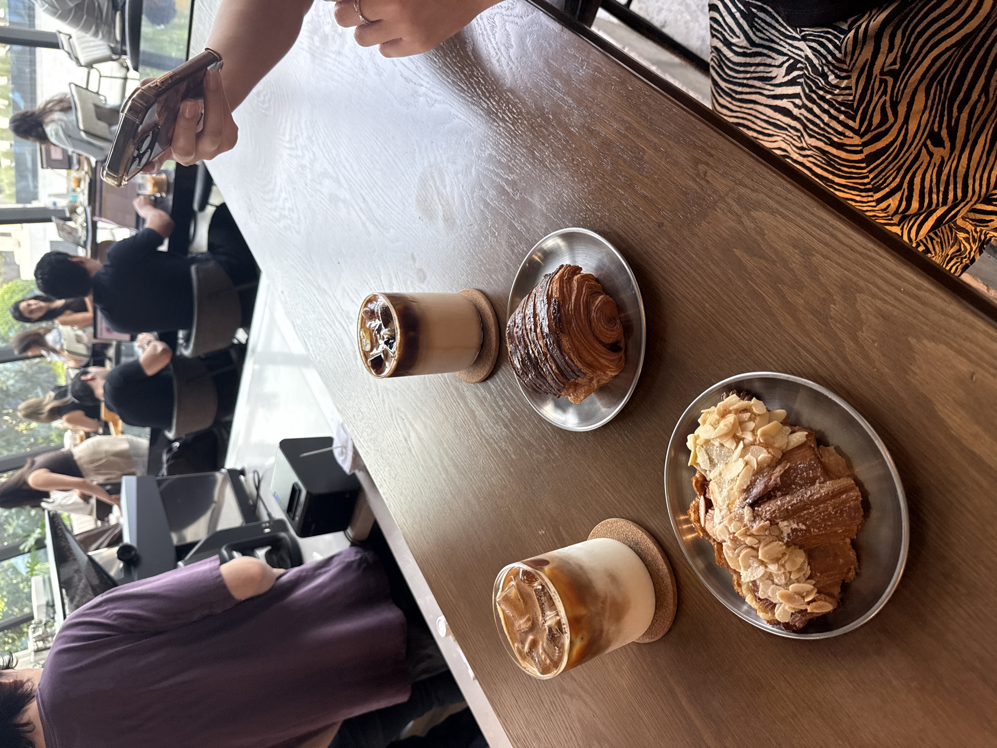 This image shows a table in a cafe in San Pedro Garza García, Mexico. Two people sit at the table, indicated by their hands, clothing, and presence in the periphery. One person wears a zebra-striped dress and holds a phone. A second person wears a purple shirt. On the table, there are two drinks in glasses on coasters and two croissants on small metal plates. Other patrons sit at tables in the background of the cafe. Sunlight filters through the large windows.