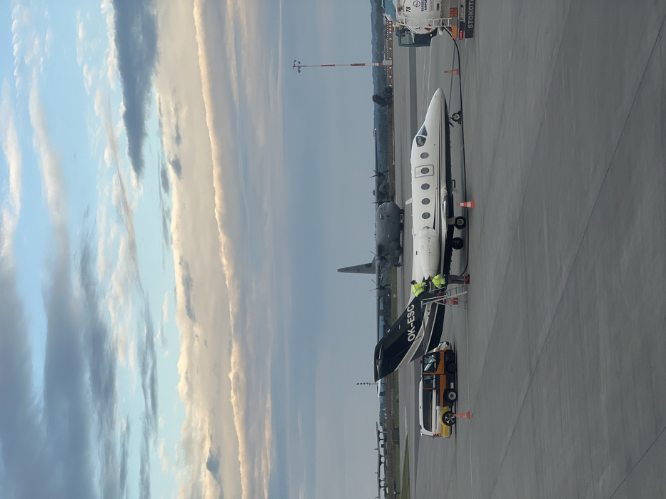 The image shows a white and black private jet, registration OK-ESC, being refueled on the tarmac of Rzeszow-Jasionka Airport in Jasionka, Poland, located at latitude 50.114 and longitude 22.026. A fuel truck is connected to the jet, and two individuals in fluorescent yellow vests are standing by the fuel truck. A white van with a red stripe is parked behind the jet, and a larger, grey military aircraft is visible in the background. The sky is cloudy, with streaks of white and grey. The tarmac is grey and appears to be clean. The image was captured during the day.
