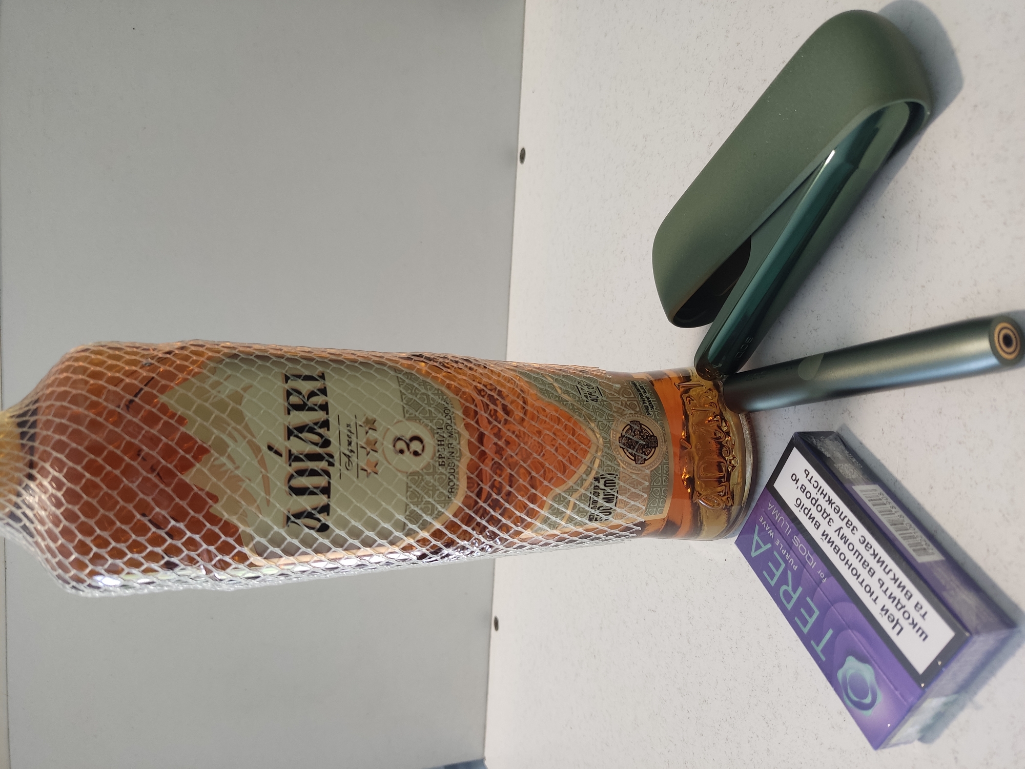 The image shows a collection of smoking-related items arranged on a white, textured surface, possibly a table or countertop. In the foreground, to the left, is a purple and white box of TEREA "Purple Wave" for IQOS ILUMA tobacco sticks. Next to the box, towards the center of the image, is a slim, metallic grey IQOS device. To the right of the device is a dark green IQOS charging case. Dominating the background is a bottle of ADJARi alcohol, wrapped in a decorative netting. The bottle appears to be a light amber color, indicating it contains a spirit like brandy or whiskey. The label on the bottle is partially visible, showing the brand name "ADJARi" and some Cyrillic text, possibly indicating origin or type. The overall setting appears to be indoors, with a plain, light-colored wall behind the objects. There are no people or animals present in the image. The lighting suggests an indoor environment with diffused light. The location is not specified in the image itself, and therefore cannot be confirmed as Radisne, Ukraine.
