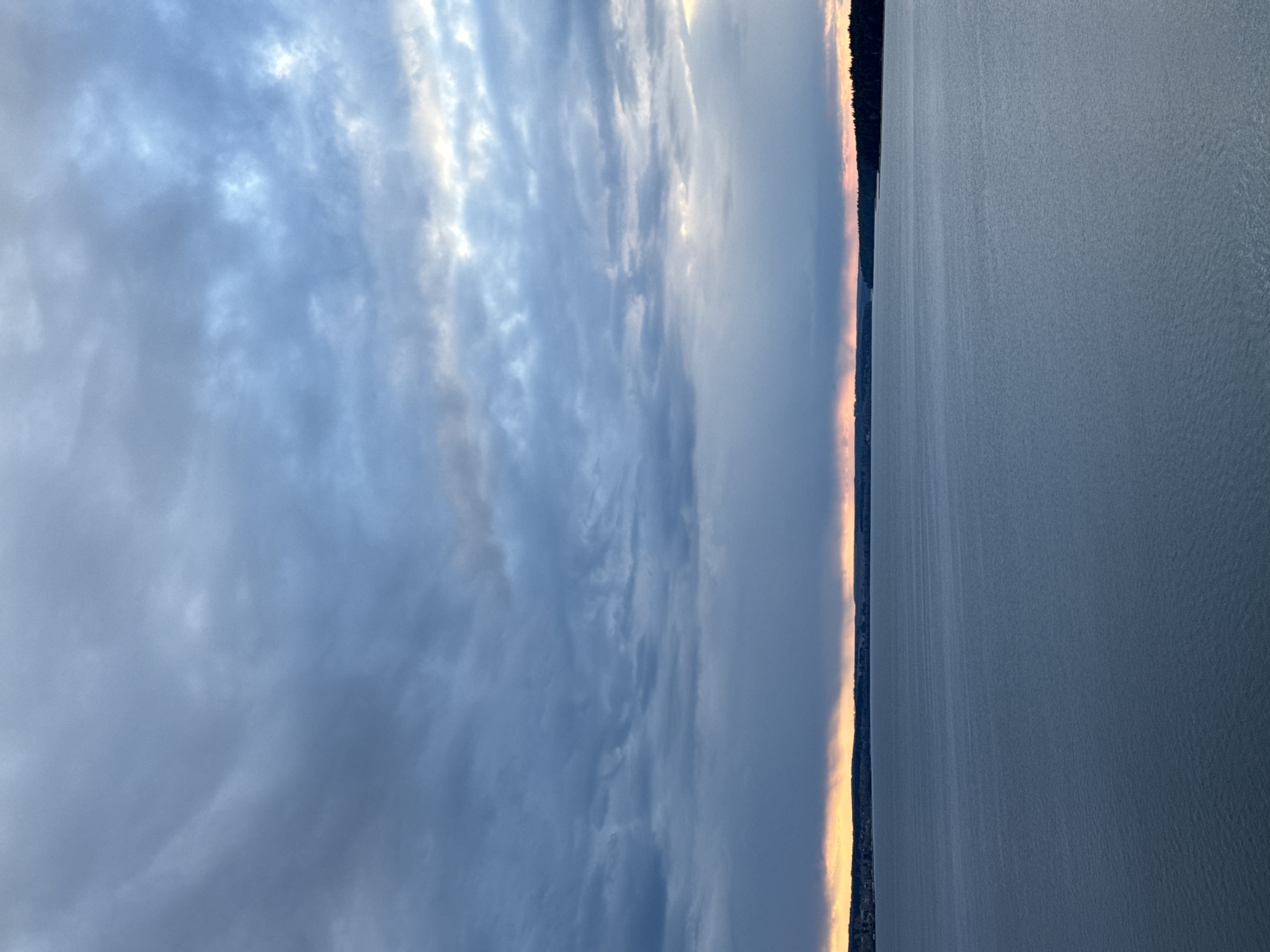 This serene landscape photograph captures a vast body of water under a dramatic, cloudy sky, likely taken at either sunrise or sunset. The horizon is marked by a distant, dark landmass, presumably covered in dense trees, separating the water from the sky. Along this horizon line, a striking band of warm, golden-orange light pierces through the clouds, indicating the low position of the sun. Above, the sky is largely overcast with soft, diffused grey-blue clouds, showing varying textures and some brighter sections where light breaks through. The extensive body of water in the foreground and mid-ground exhibits a subtly rippled surface, reflecting the muted colors of the sky. No individuals, specific activities, or man-made structures are visible, emphasizing the natural tranquility and grandeur of the scene. The weather appears to be overcast with a breaking sky, creating a soft, diffused lighting environment. Consistent with the provided context, this is likely a view of the Votkinsk Reservoir in Votkinsk, Russia. There is no visible text in the image.