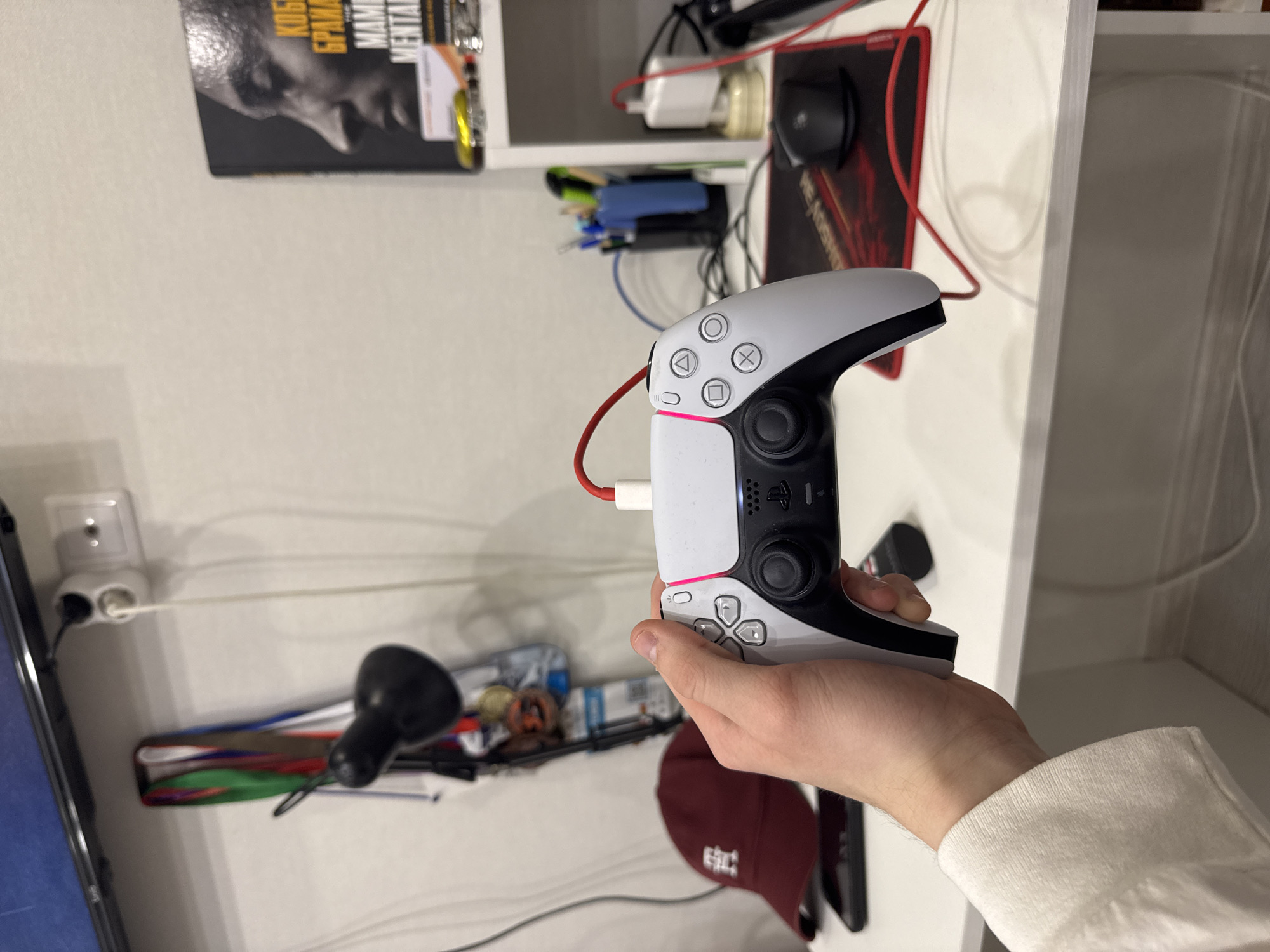 The image depicts an indoor scene, likely a personal room in Kazan, Russia, focusing on a person's hand holding a PlayStation 5 DualSense controller. The controller, predominantly white with black accents, is connected by a red USB-C cable, and a pink/red light strip is illuminated on its front. The person's hand, fair-skinned and wearing a light-colored long-sleeved top, holds the controller in the foreground.
In the background, white wall-mounted shelves are visible, cluttered with various items. On the top shelf, a book with the partial title "KOBE БРАЙНТ MENTAL" (suggesting "Kobe Bryant: The Mamba Mentality") is prominent, alongside a yellow trinket, a white power adapter, a black gaming mouse resting on a red and black "REDRAGON" mousepad, and a tangle of wires. The middle shelf contains more cables and what appears to be writing instruments in a blue holder. A lower shelf or surface shows a partial view of a red baseball cap and other indistinct items.
A European-style double power outlet is visible on the left wall, with several cables plugged in and running along the white wall. A black desk lamp head is partially visible on a surface below the shelves, and a screen or monitor is cropped on the far left. The scene suggests a casual, lived-in space with a focus on gaming and personal electronics. No specific time of day or weather is discernible.