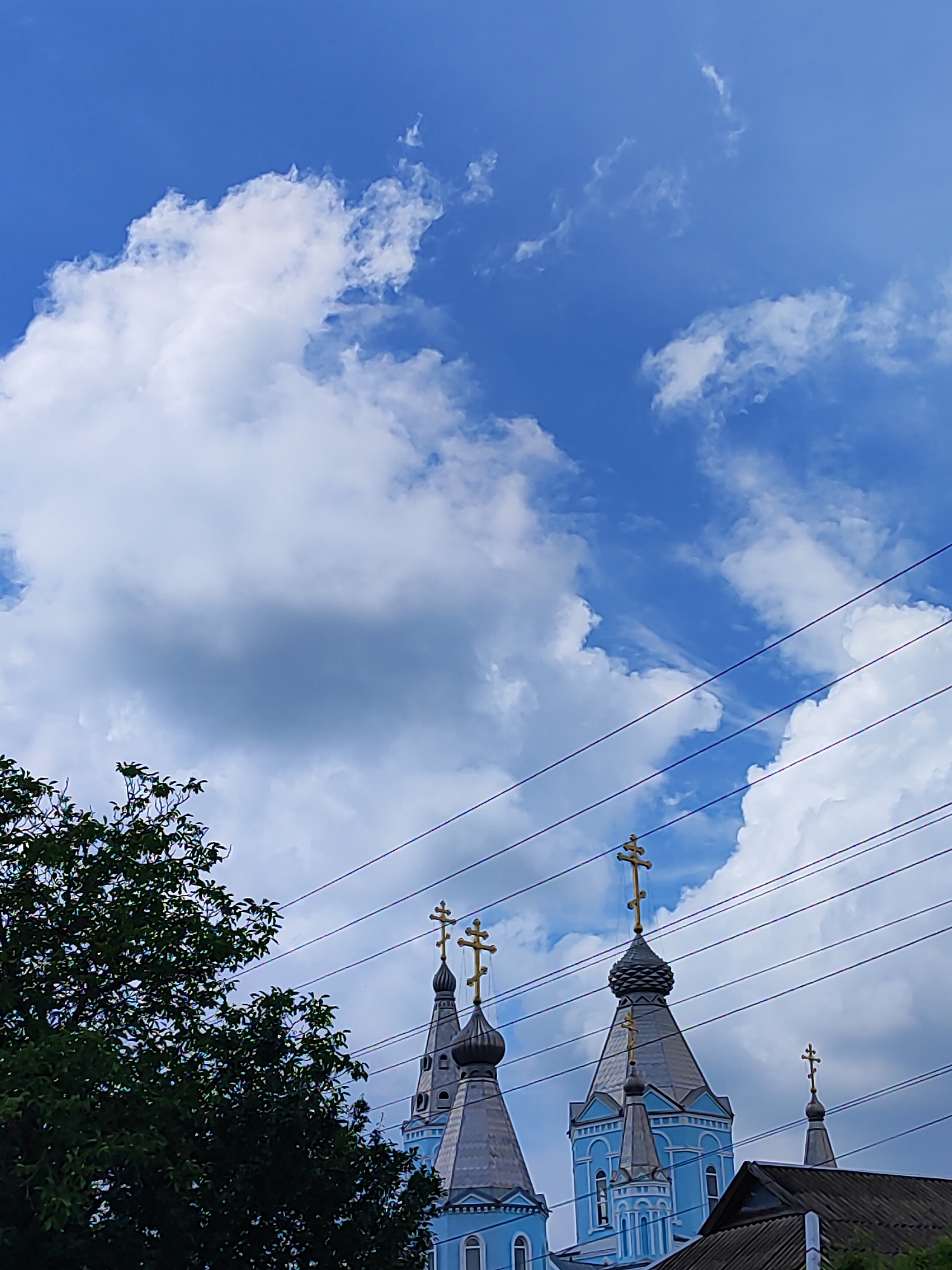 The image shows the domes of a blue church in Bryukhovetskaya, Russia. The domes are topped with golden crosses. A large green tree is visible on the left side of the image. Power lines are strung across the sky, which is blue with white clouds. The bottom right corner of the image shows the roof of another building.