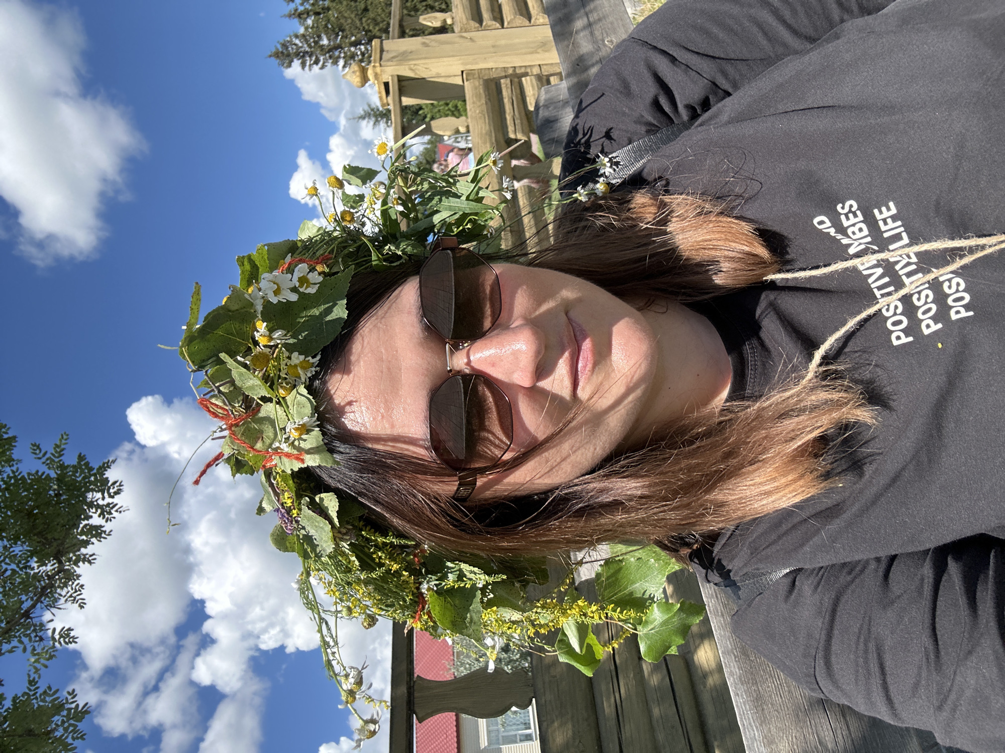 A woman wearing a flower crown and sunglasses poses for a selfie outdoors on a sunny day. She is sitting on a wooden bench, with a rustic wooden fence behind her. In the background, a building with a red roof and a tall wooden structure are visible. The sky is blue with scattered white clouds. The woman is wearing a black t-shirt with white text that reads "POSITIVE MIND POSITIVE VIBES POSITIVE LIFE". The location is indicated as the city of Bolsherechye, Russia.