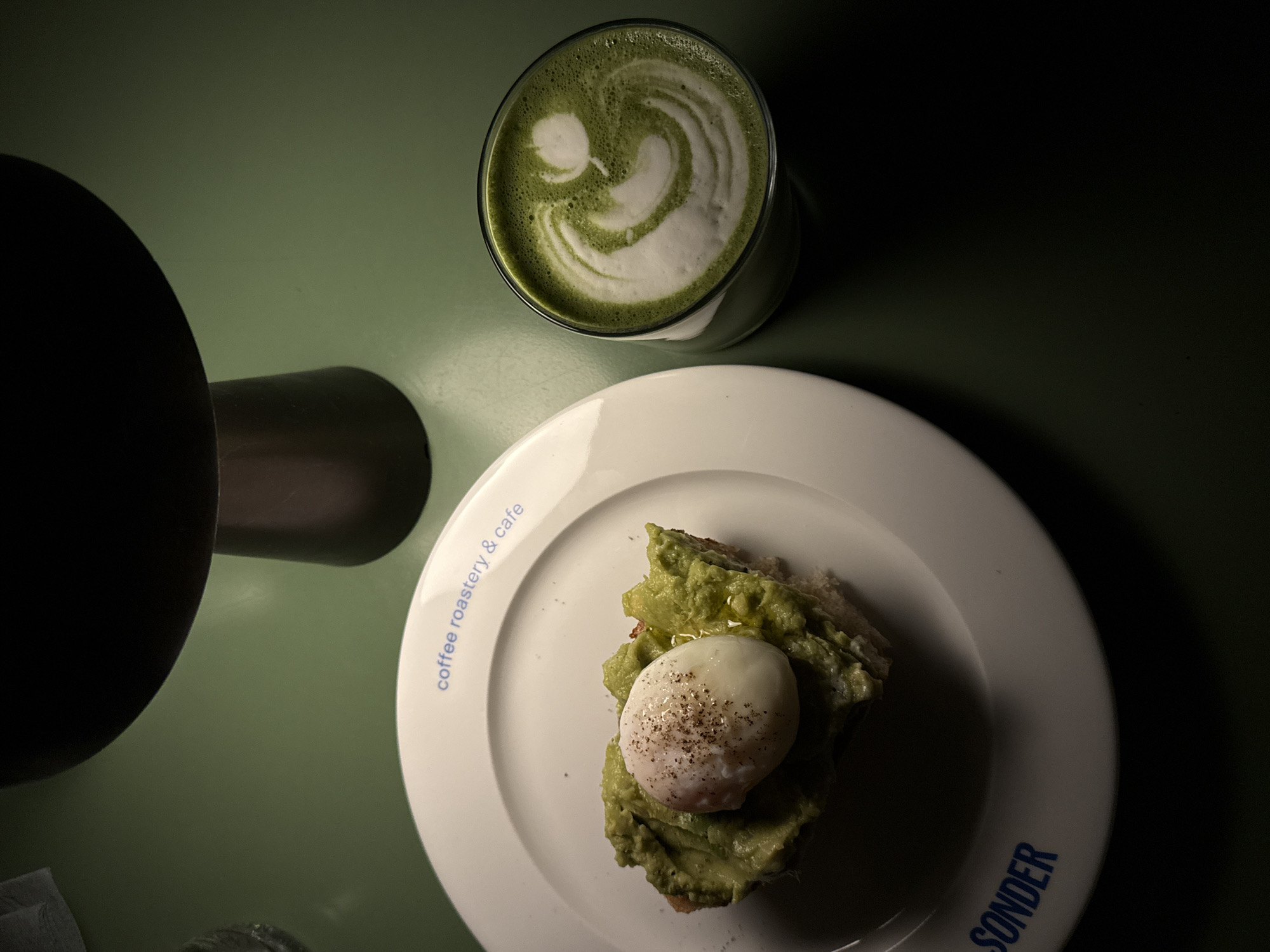 A top-down, slightly angled view captures a table setting with a glass of matcha latte, a plate of avocado toast topped with a poached egg, and part of a lamp. The latte has a white, swirled foam design on top. The avocado toast is on a white plate with the words "coffee roastery & cafe" and "SONDER" printed in blue. The table surface is a muted green. The scene appears to be indoors, illuminated by a lamp whose base and shade are partially visible on the left. Based on the plate, the location is most likely the Sonder coffee roastery & cafe, which has a branch in Belgrade, Serbia.
