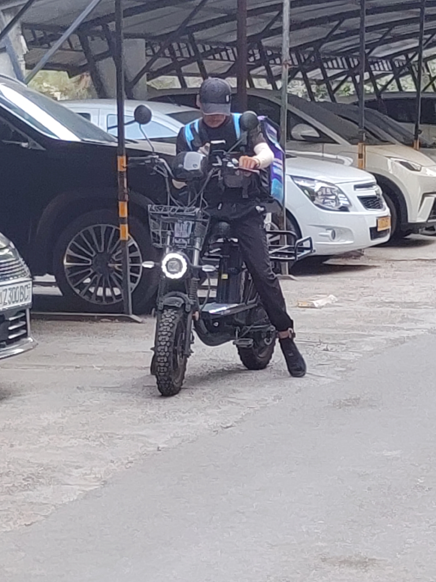 A person in a black baseball cap and a blue and black uniform is seated on a black electric scooter with a basket in front. The person is holding a mobile device in their left hand and appears to be operating the scooter. The scooter has a circular headlight. The person is wearing black pants and dark shoes. The scooter is parked on a paved surface, possibly a parking lot, in front of several parked cars. The background shows a roofed structure providing shade, and the overall environment suggests an outdoor urban setting in Tashkent, Uzbekistan. The license plate of a silver car on the left is partially visible and reads "UZ 300 BC".