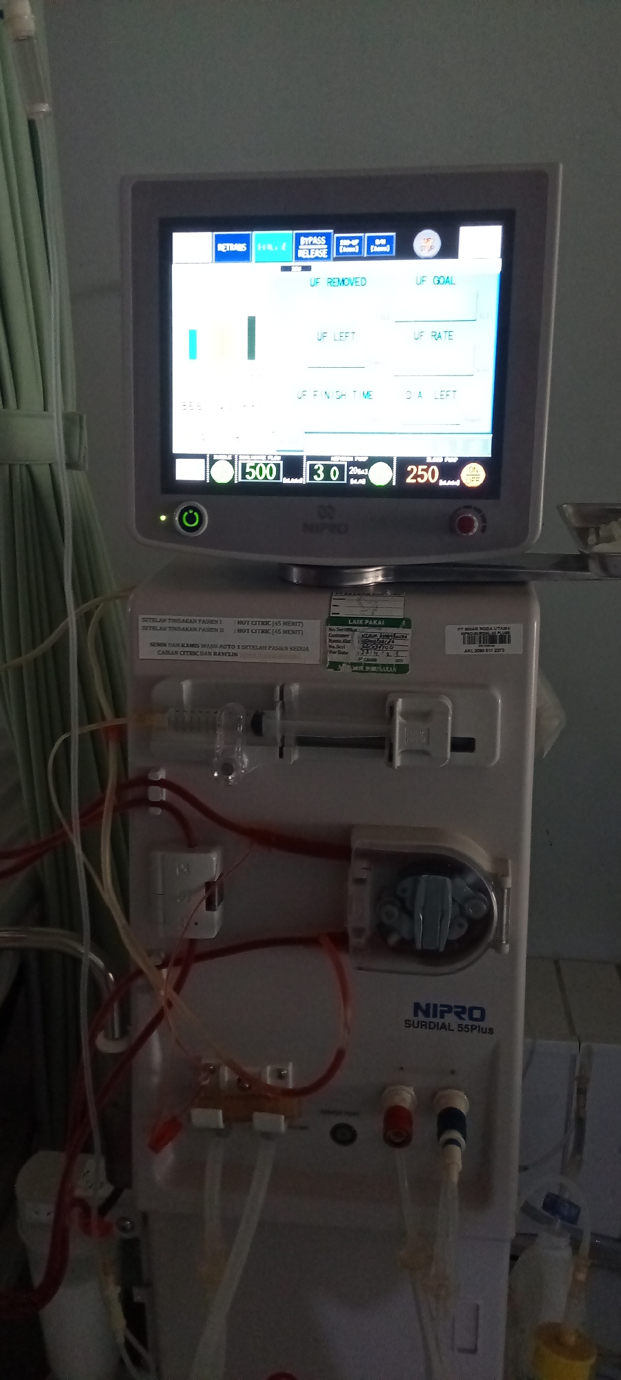 The image captures a Nipro Surdial 55Plus hemodialysis machine in an indoor medical setting, likely a hospital or clinic in Lhokseumawe, Indonesia. No people are visible.
The machine's screen is illuminated, displaying various parameters crucial for a dialysis treatment. Key metrics visible include "UF REMOVED," "UF GOAL," "UF LEFT," "UF RATE," and "UF FINISH TIME," all related to ultrafiltration. Numerical readings show "500" (likely blood flow in mL/min), "30" (possibly arterial or venous pressure), and "250" (likely dialysate flow in mL/min). Buttons on the screen include "RETRANS," "BYPASS," "RELEASE," and "STOP." A green power indicator light on the machine's front indicates it is powered on.
Below the screen, medical tubing, some containing red fluid suggestive of blood, is connected throughout the machine. A syringe pump and a roller pump, essential components for blood circulation during dialysis, are also visible. A green curtain hangs to the left of the machine.
Visible text on labels and stickers on the machine includes maintenance instructions in Indonesian: "SETELAH TINDAKAN PASIEN I : HOT CITRIC (45 MENIT)" (After patient action I: HOT CITRIC (45 MINUTES)) and "SETELAH TINDAKAN PASIEN II : HOT CITRIC (45 MENIT)" (After patient action II: HOT CITRIC (45 MINUTES)). Another instruction reads: "SENIN DAN KAMIS WAJIB AUTO 2 SETELAH PASIEN KEDUA CAIRAN CITRIC DAN RAYCLIN" (Monday and Thursday mandatory auto 2 after second patient CITRIC and RAYCLIN fluid). A "LAIK PAKAI" (Fit for use) label is present, along with a partial "Expired Date" (appearing as "23/..."), and a product registration number "AKL 2080 511 2373" from "PT BHAN RODA UTAMA." The machine's brand, "NIPRO SURDIAL 55Plus," is clearly visible on its body.