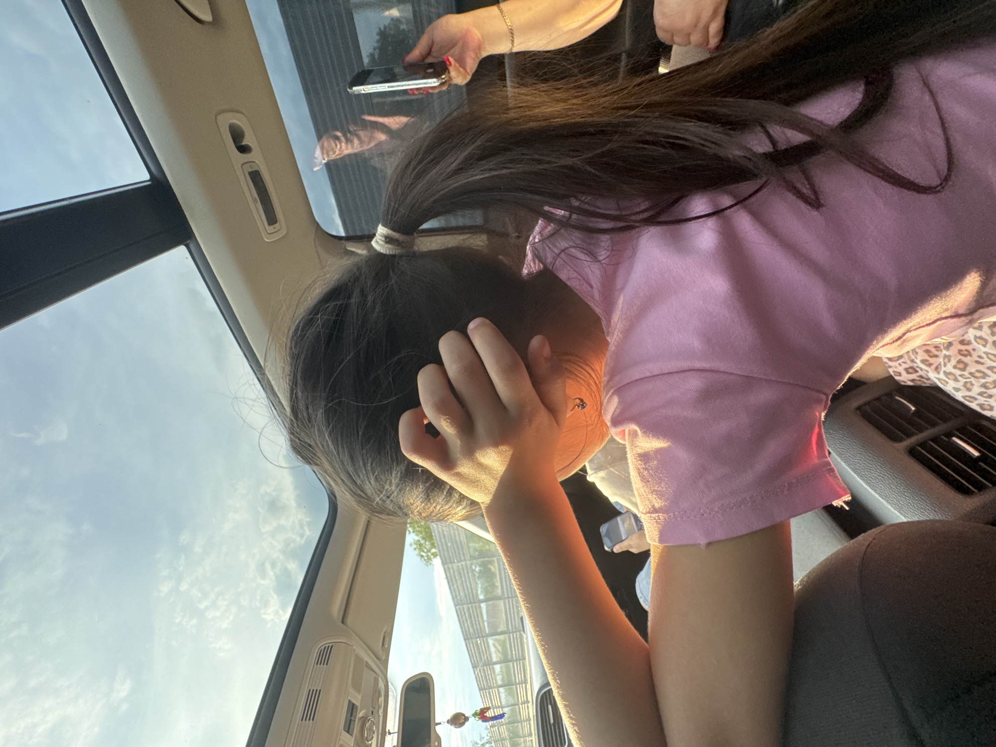 The image depicts the interior of a vehicle, likely a car or SUV, in motion on a highway or road in Suprunivka, Ukraine. A panoramic sunroof reveals a clear blue sky with scattered white clouds overhead. Direct sunlight, indicative of golden hour, illuminates the scene from the left, casting warm light and shadows.
In the foreground, a young person, possibly a pre-teen female, is seated. She has long, dark hair tied into a ponytail with a light-colored elastic. She is wearing a pink short-sleeved top and leopard-print patterned lower garments. Her right arm is raised, with her hand partially covering her face or head, possibly shielding from the sun or in a contemplative pose. A delicate necklace with a small pendant is visible around her neck.
To the right of the frame, partially visible, is the right arm and hand of another individual, likely an adult. This hand, adorned with a thin gold chain bracelet and red nail polish, holds a silver-colored smartphone vertically, pointing towards the vehicle's side window, suggesting the act of taking a photo or video.
The vehicle's interior features a light-colored headliner with various controls and air vents, a rearview mirror with a hanging charm, and rear cabin air vents on the console. Outside the windows, a long grey sound barrier or highway fence structure is visible parallel to the road, with green foliage visible beyond it. A hand holding another smartphone is partially visible in the lower-left, possibly belonging to the foreground individual or another passenger.