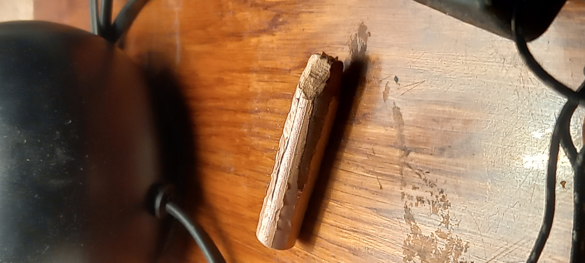 The media file presents a close-up, overhead view of a light brown, cylindrical object, likely a piece of wood, resting on a wooden surface. The object, resembling a short stick or dowel, exhibits a rough, chipped texture, with visible wood grain and variations in color from light tan to darker brown.
The surface it rests upon is a polished wooden tabletop or floor, characterized by a warm, reddish-brown hue. This surface shows signs of wear, including numerous scratches, scuffs, and possibly some residue or discoloration, suggesting age or frequent use.
In the background, on the left side of the frame, a large, dark, curved object is partially visible, possibly part of a lamp base, a piece of furniture, or an appliance. A thin, black cable extends from the lower left corner towards the right, appearing to be connected to or associated with the dark object.
The scene is static, depicting no active event or people. The lighting suggests an indoor setting, possibly under natural light or a bright artificial source, highlighting the textures of the wood. No visible text is present in the image. This detailed shot captures a mundane object within an everyday setting in Kryvyi Rih, Ukraine.