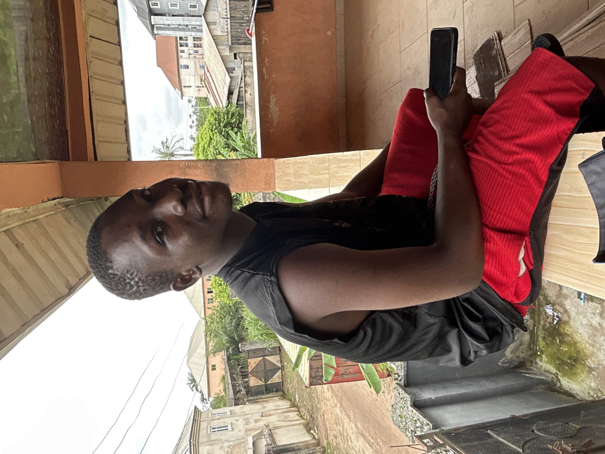 The image shows a young Black man sitting outdoors, holding a smartphone. He is wearing a black sleeveless shirt and red shorts with a ribbed texture. The man is looking towards the camera with a relaxed expression.
The setting appears to be a residential area with buildings in the background. There are indications of vegetation, including trees and plants. The ground is tiled in some areas and appears to be dirt or gravel in others. A set of stairs is visible in the lower right portion of the image. The overall lighting suggests it is daytime, possibly overcast given the bright but diffused light.
The man is holding the smartphone in his right hand, and the device is oriented vertically. The image is taken from a slightly elevated angle, looking down at the man. There is some graffiti or writing on a wall in the background, with the word "DANNYIE" partially visible. The context provided indicates the location is Ihiagwa, Nigeria.