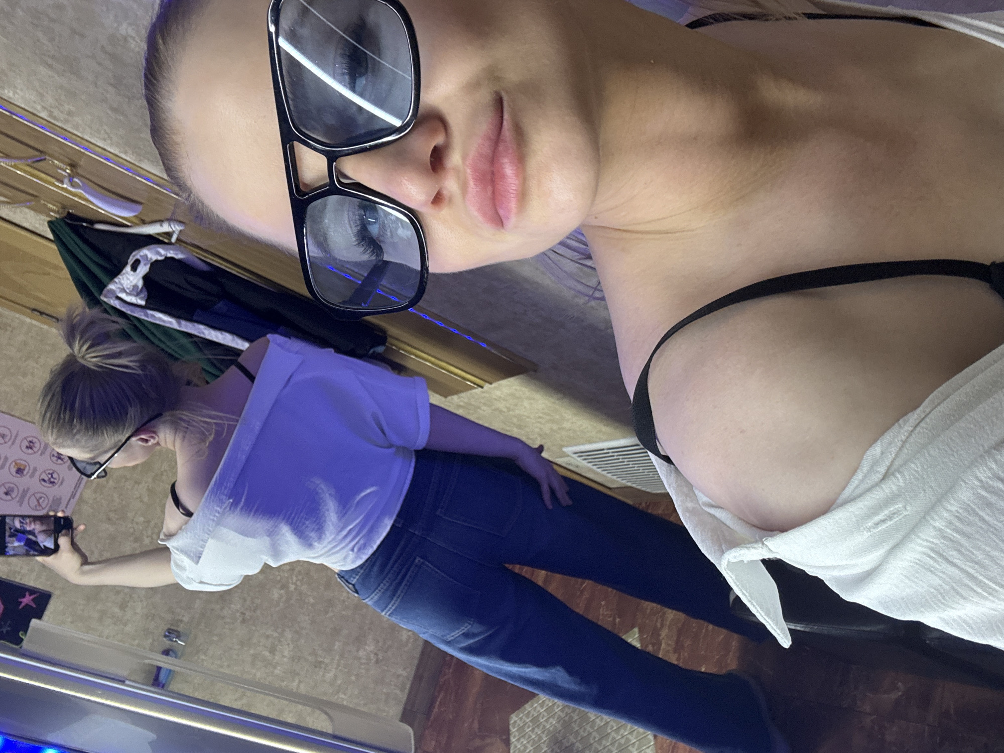 The image shows a fair-skinned woman wearing black-framed sunglasses and a white top with a black bra strap visible. She is indoors, presumably in a tanning salon, as a tanning bed can be seen in the background. The woman is taking a selfie, with another woman visible in the mirror holding a phone and wearing a white top and jeans. A poster with tanning instructions is visible on the wall. The location is unknown.