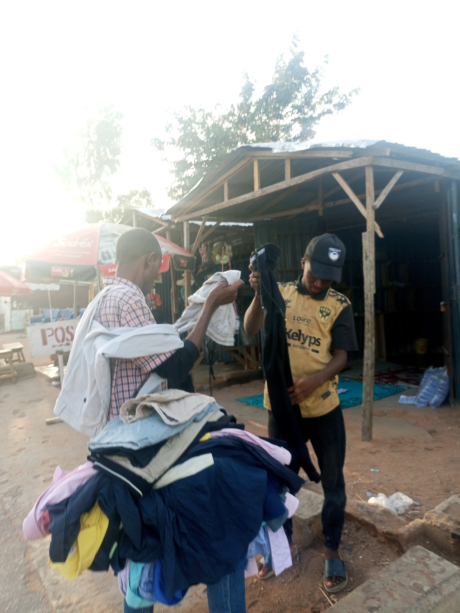 This outdoor image, taken in Tempure, Nigeria, depicts two men actively involved in sorting or displaying clothing for sale under bright, sun-drenched conditions, likely morning or late afternoon.
In the foreground, a substantial pile of folded clothes, including shirts and trousers in various colors such as pink, yellow, blue, black, and beige, is prominently displayed, suggesting merchandise.
To the left, a man wearing a plaid or checkered long-sleeved shirt holds up a light-colored item of clothing, possibly a shirt or jacket. A light-colored jacket is draped over his left shoulder. Behind him, parts of a market area are visible, including a red umbrella with "Sudrex" printed on it and a partially visible sign that reads "POS".
To the right, another man, wearing a black baseball cap and a yellow and black short-sleeved jersey, is holding up a dark, possibly black, piece of clothing. The jersey clearly shows "Loire" and "Kelyps INTERIM" in black text. He is wearing dark trousers and dark sandals. Behind him, a rustic market stall with a wooden frame and a corrugated metal roof stands, its interior dark. A light blue mat and some bundled items are on the ground inside the stall.
The ground is unpaved dirt, scattered with rocks and debris. The sky in the upper left portion of the image is heavily overexposed due to direct sunlight. Trees are visible against the bright sky in the background. The scene conveys a sense of everyday market activity in a sunny, outdoor setting.