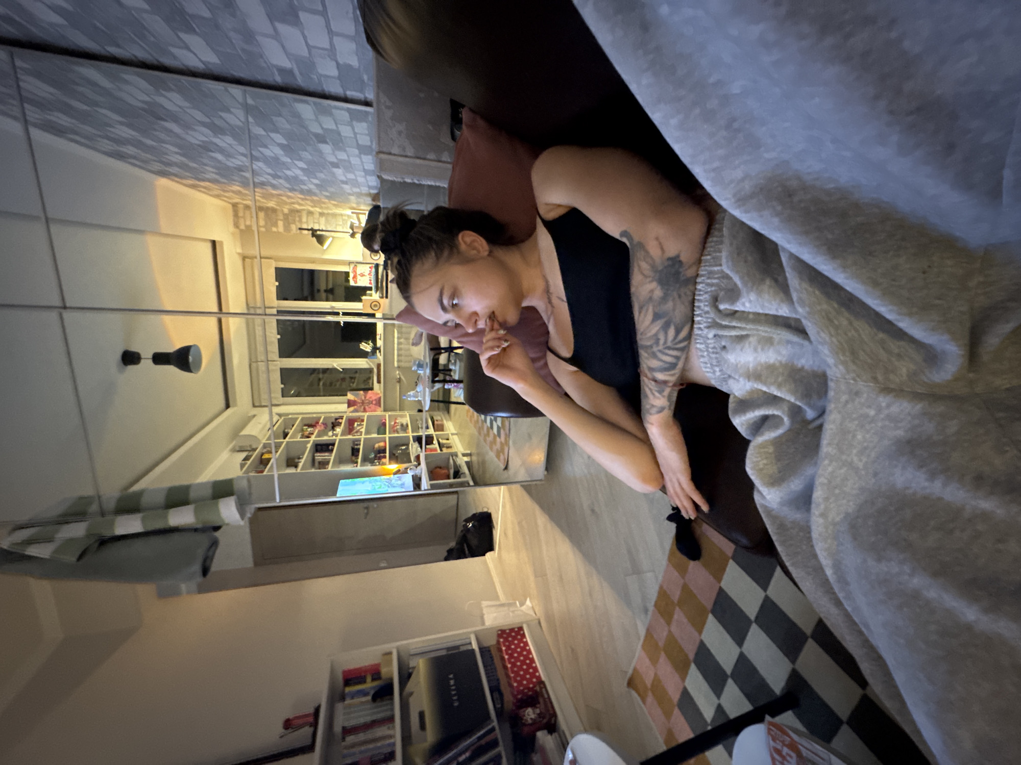A young woman with dark hair tied up in a bun is relaxing on a couch in a modern apartment, possibly located in Moscow, Russia. She is wearing a black tank top and grey sweatpants, with visible tattoos on her arms. The room features a large mirror, reflecting a view of shelves filled with books and decorative items, as well as a window showing a city at night. The overall ambiance suggests a cozy evening at home.