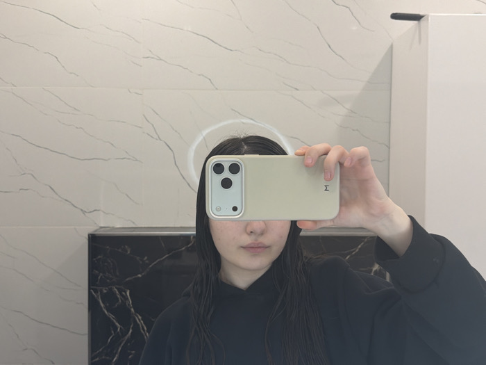 A person with dark hair is holding a smartphone up to their face, obscuring their eyes, likely taking a selfie in a bathroom. The phone has a light-colored case. Behind the person is a wall with white marble-like tiles and a black marble countertop or cabinet. A mirror is visible reflecting the person. A white ring reflection surrounds the person's head. The person is wearing a black hoodie.