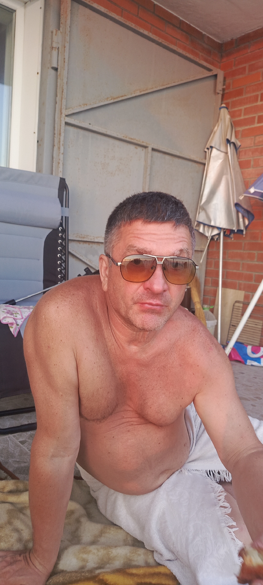 A man is sitting on a towel outdoors, wearing sunglasses and wrapped in a white towel. He is partially shirtless, with his torso and arms visible. He appears to be in a relaxed, casual pose. In the background, there is a brick wall, a metal structure, and a folded umbrella. The setting suggests a warm, possibly summer day. The man's location in Bolshoy Kamen, Russia is not discernible from the visual information in the image.