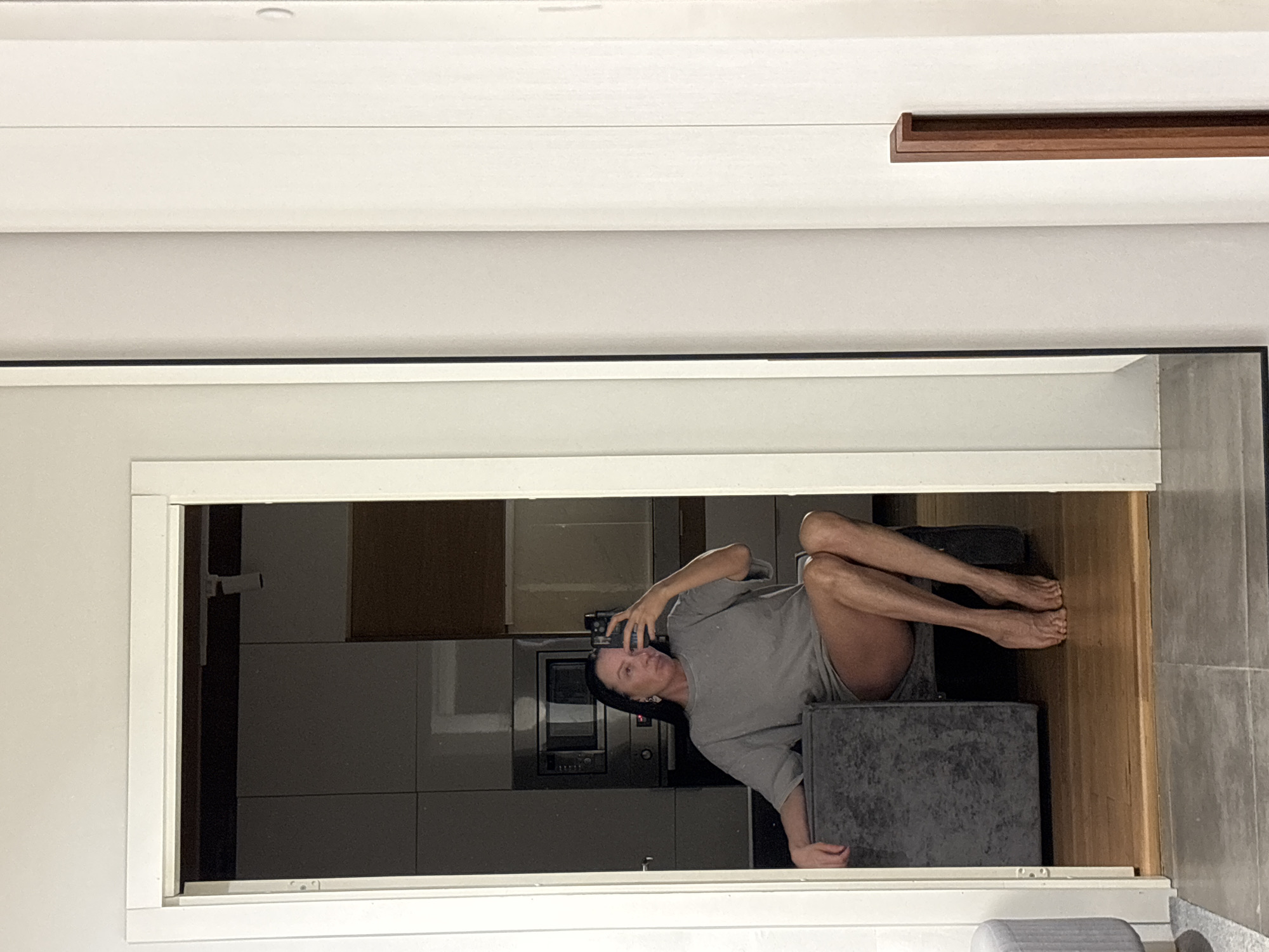 The image displays a mirror selfie of an adult female situated indoors. The subject has long dark hair and is wearing a loose, grey short-sleeved t-shirt, exposing her bare legs and feet. She is seated on a dark grey upholstered chair with her left arm resting on its armrest. Her legs are crossed at the ankles, and her bare feet are visible on a light brown wooden floor. She is wearing earrings and holds a black smartphone in her right hand, angled to capture her reflection.
The background visible in the mirror's reflection depicts a modern residential kitchen or living space. It includes dark grey flat-panel cabinets, a built-in stainless steel microwave oven, and an oven below it. Some lighter-colored wall cabinets and a dark brown accent strip are visible in the upper right portion of the reflection. Overhead track lighting is present in the reflected room.
The mirror, framed by a thin dark border, stands vertically in a white-framed doorway, resting on a grey tiled floor. To the immediate left of the mirror, a portion of a light grey upholstered ottoman is visible. To the right of the mirror, a vertical dark wooden handle or decorative strip is affixed to a light-colored wall or furniture. The specific location within the city of Chaikovsky, Russia, is not discernible from the visual evidence.
