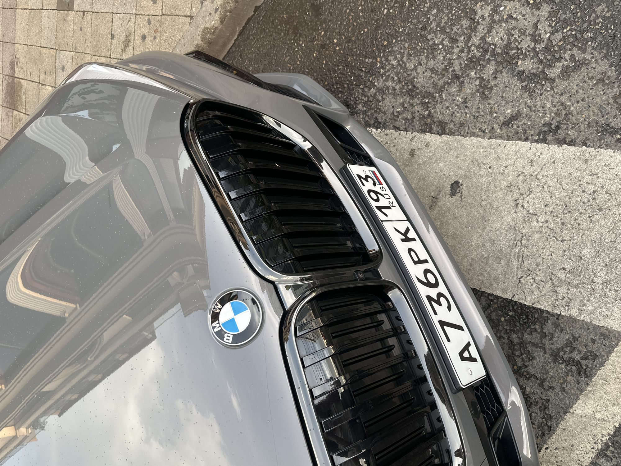 A close-up, angled shot shows the front grille and hood of a gray BMW. The car's hood reflects the surroundings, appearing slightly wet with small water droplets. The iconic BMW logo, a blue and white circular emblem, is visible on the left side of the grille. The car has a black, slatted grille with chrome accents. A Russian license plate, reading "A736PK 193 RUS", is mounted on the front. The license plate has a white background with black lettering and a small Russian flag symbol.
The car is parked on a surface that appears to be a paved area with some texture, possibly a street or sidewalk. The ground is a mix of gray concrete pavers and darker asphalt, with a white line painted on the ground. The lighting suggests it is daytime, with shadows cast by the car and possibly buildings or trees. The weather seems partly cloudy, as indicated by the reflections on the car's hood. The scene appears to be a typical urban or suburban setting in Krasnodar, Russia. No people are visible in the image.