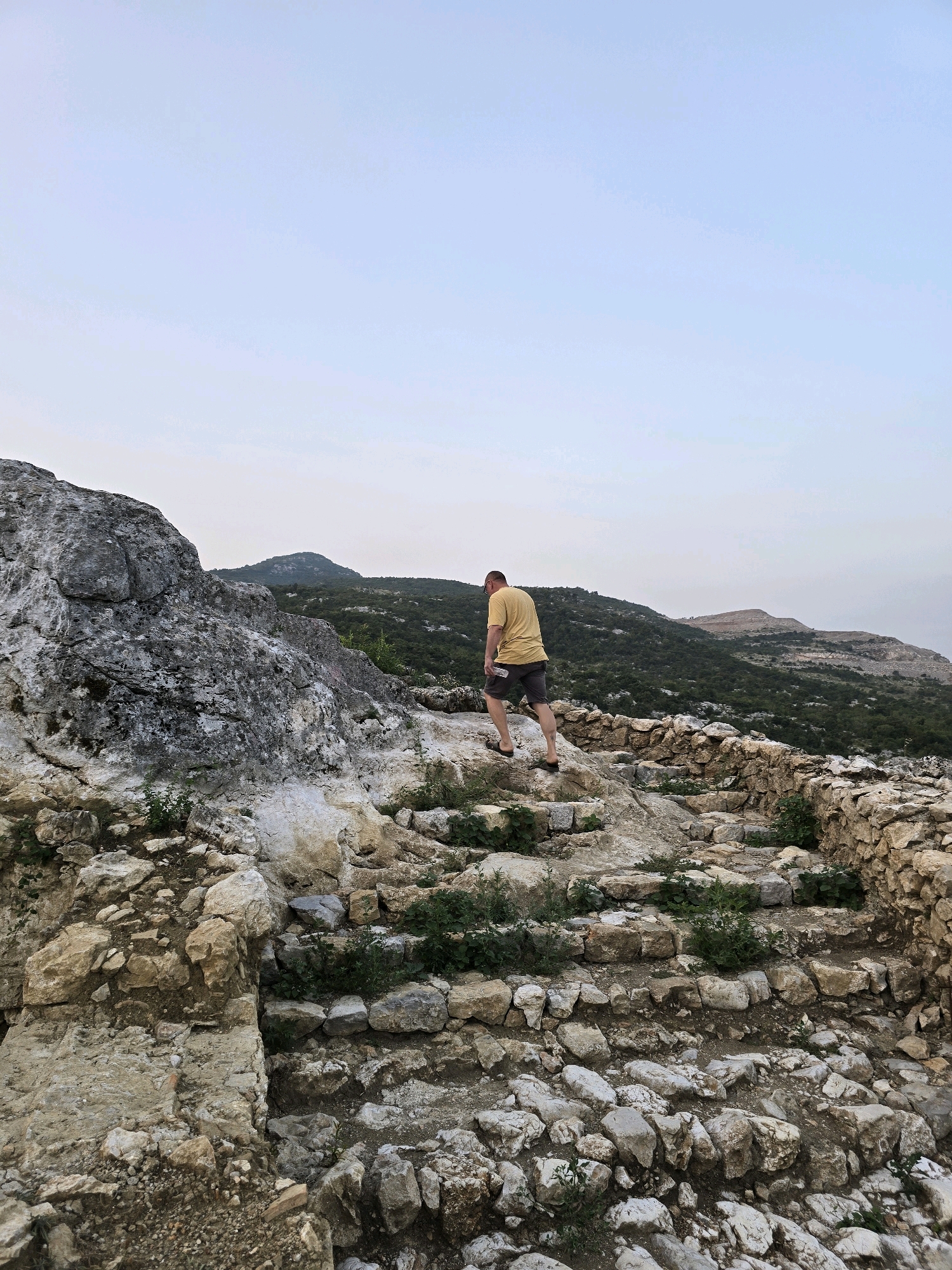In the city of Gornja Vrbica, Montenegro, a man in a yellow t-shirt and grey shorts walks up a weathered stone staircase. The staircase is integrated into a rocky, uneven terrain, with remnants of a stone wall on the right. Greenery sprouts between the stones, indicating an outdoor, natural environment. In the background, rolling hills covered in dense green vegetation stretch towards a pale, overcast sky. The overall scene depicts an archaeological site or a hiking path in a mountainous region.