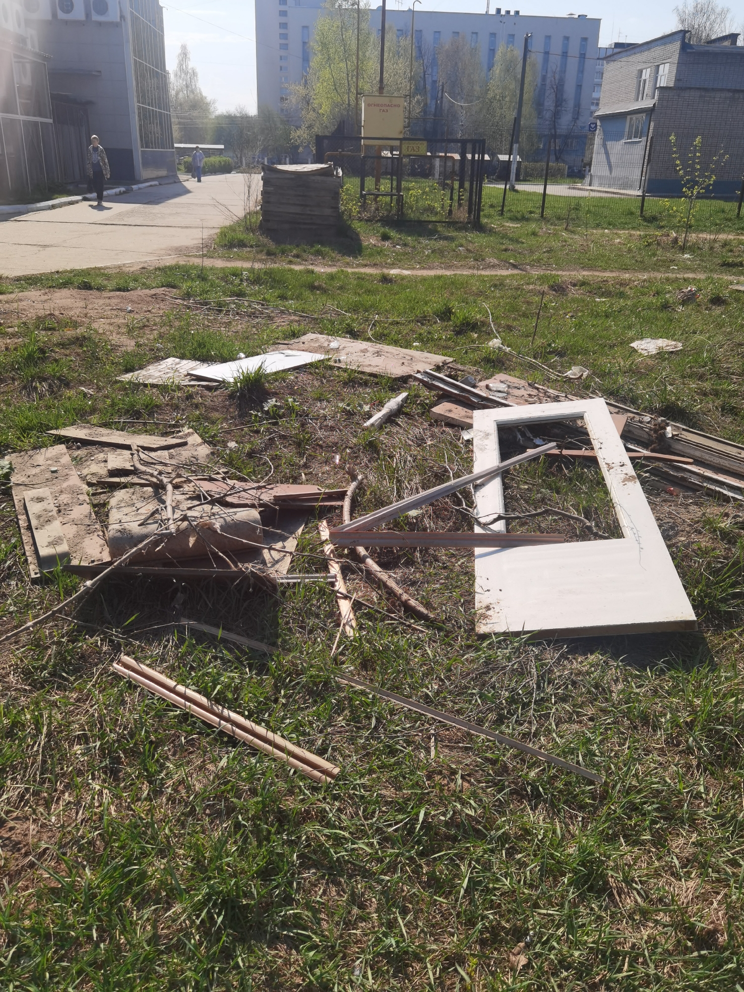 The image depicts a debris field in a grassy area, likely in the city of Chaikovsky, Russia. The debris includes pieces of wood, a door frame, and other construction materials. In the background, there are buildings and a paved walkway with people walking on it. The area appears to be an open space near buildings, with a stack of wood pallets and fencing in the distance. The scene suggests illegal dumping or improper disposal of construction waste.