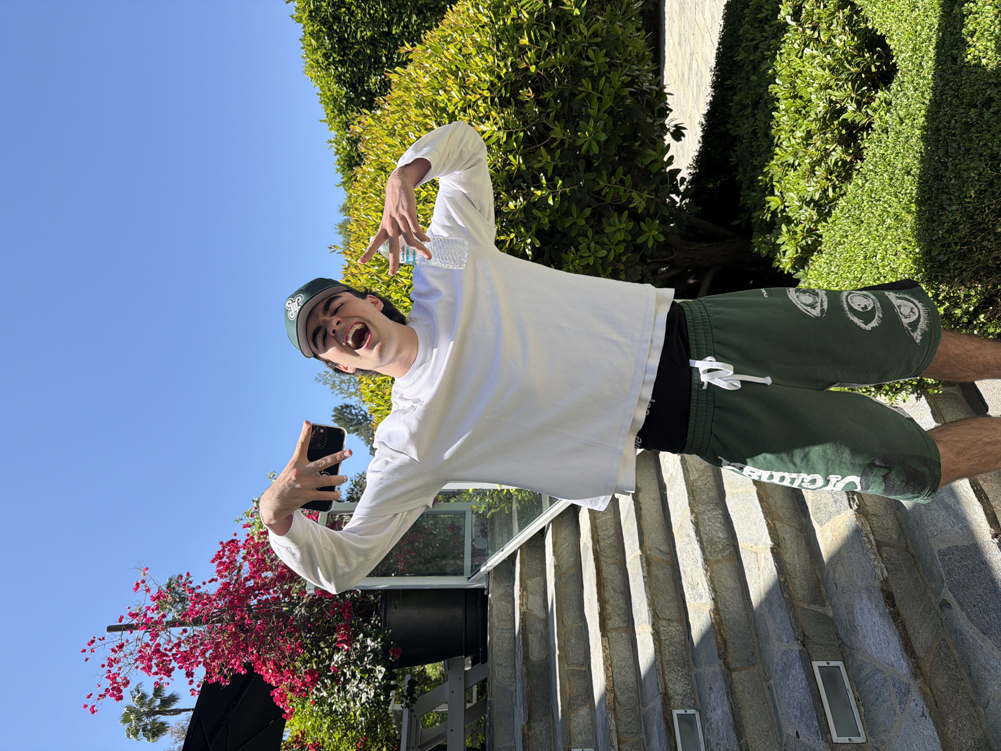A young man in a white long-sleeved shirt, green shorts, and a green baseball cap is taking a selfie with a smartphone on stone steps in Los Angeles, California. He is smiling widely and making a hand gesture. The background features lush greenery and a blue sky. A bougainvillea is blooming near the stairs.