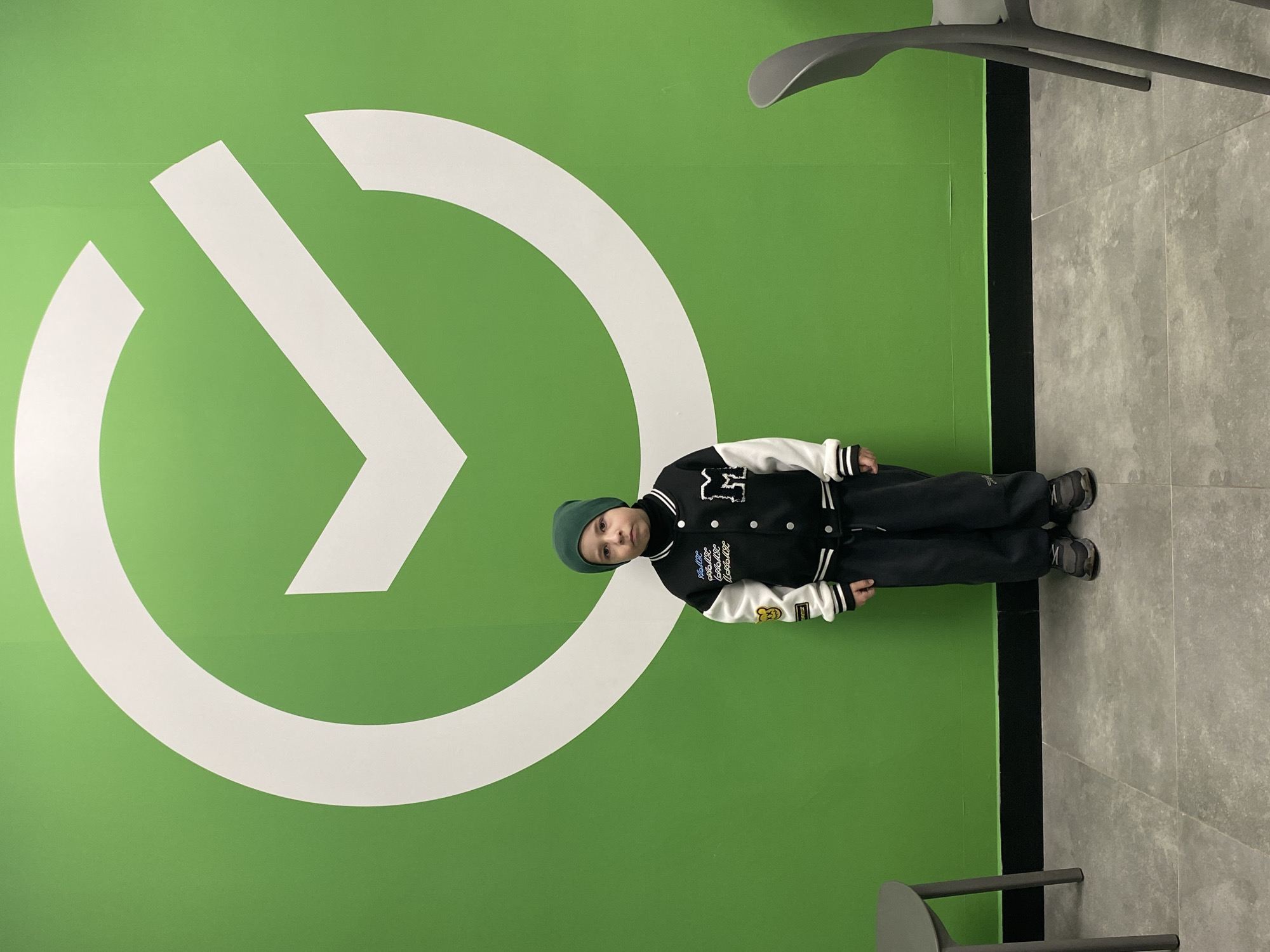 A child wearing a dark green beanie, a black and white varsity jacket, and dark pants is standing in front of a bright green wall with a large white logo on it. The child is facing slightly towards the right of the frame. The background includes a dark gray tiled floor and some gray chairs. The location is Марковский, Russia.