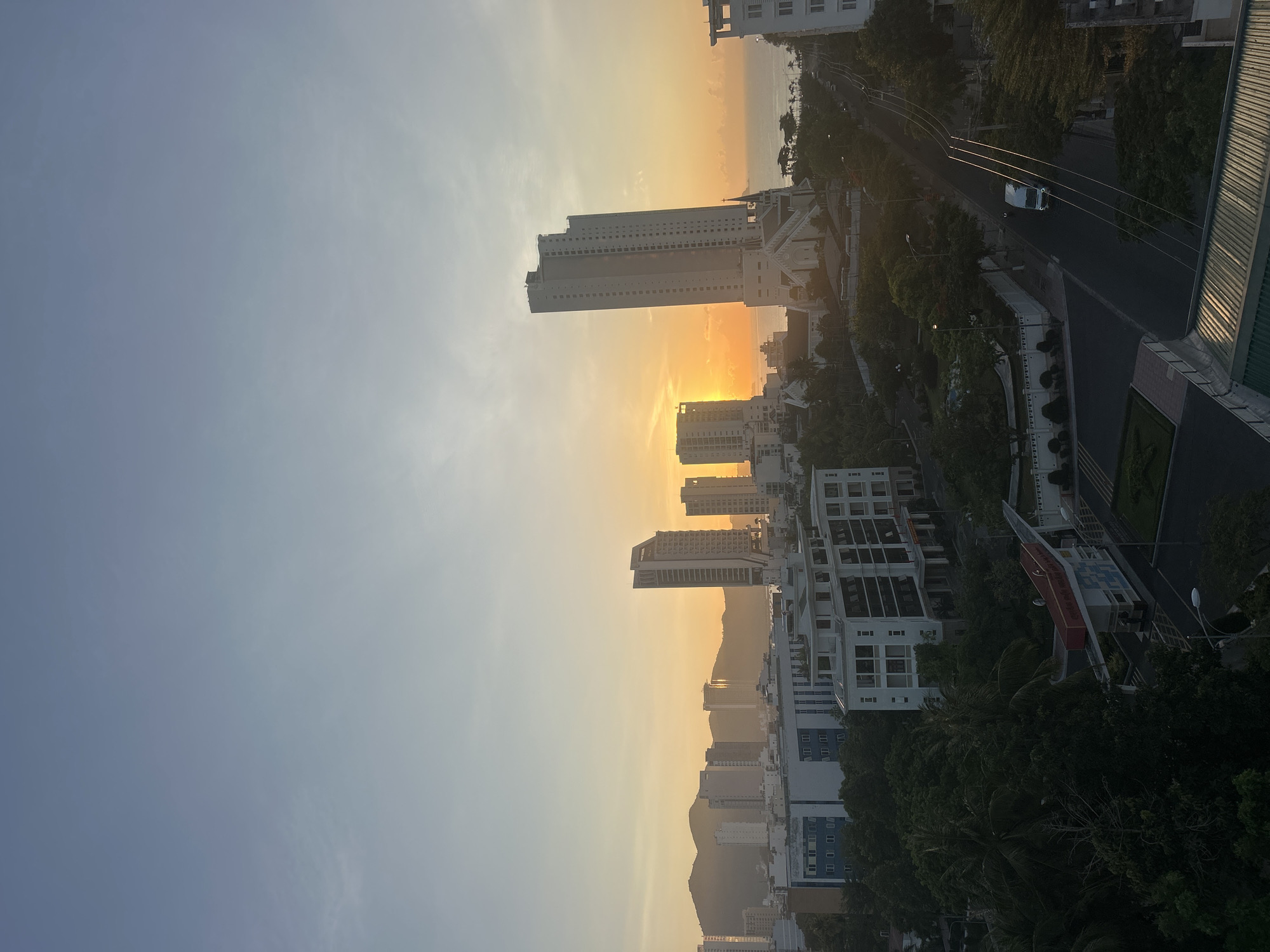 The image captures a cityscape at sunset in Bac Nha Trang Ward, Vietnam. Tall buildings, silhouetted against the golden sky, dominate the view. Some buildings are high-rise structures with many floors, while others are shorter and more spread out. Mountains are visible in the distance, creating a backdrop to the city. The sky is gradient, transitioning from pale blue to orange and yellow near the horizon where the sun is setting. In the foreground, there are trees and a road, giving a sense of depth to the scene. A white car is seen on the road. The overall atmosphere is one of peace and tranquility.