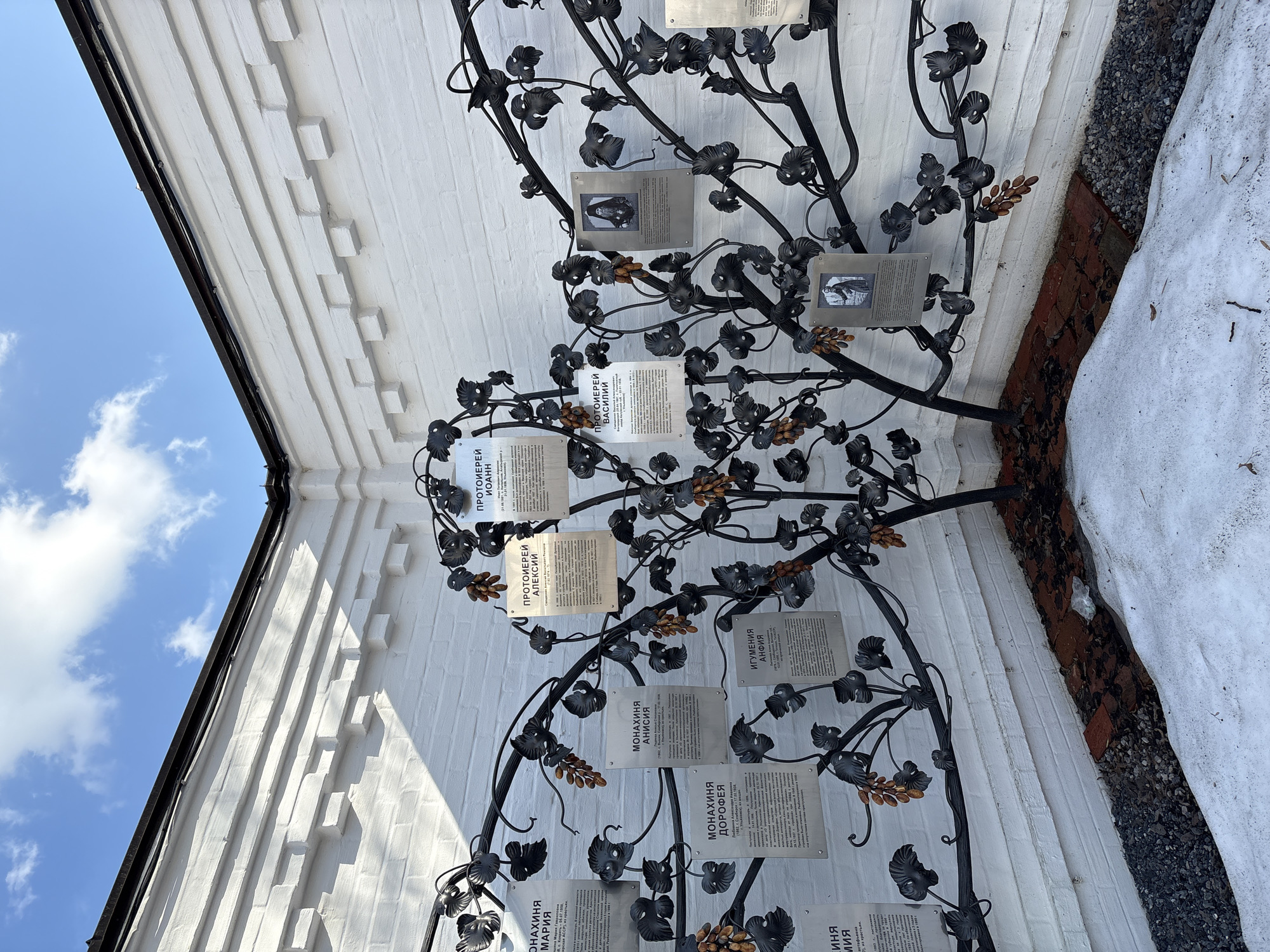 The image shows a metal tree sculpture affixed to a white brick wall. The tree is adorned with metal leaves and grapes, along with plaques that contain inscriptions in Russian. Some plaques have photographs. The inscription "МОНАХИНЯ МАРИЯ" is readable on one plaque, translating to "Nun Maria." Other readable inscriptions include "МОНАХИНЯ ДОРОФЕЯ" (Nun Dorothea), "МОНАХИНЯ АНИСИЯ" (Nun Anisia), "ИГУМЕНИЯ АНФИЯ" (Abbess Anfia), "ПРОТОИЕРЕЙ АЛЕКСИЙ" (Archpriest Alexei), "ПРОТОИЕРЕЙ ИОАНН" (Archpriest Ioann), and "ПРОТОИЕРЕЙ ВАСИЛИЙ" (Archpriest Vasilii).
To the right of the tree's base is snow and a patch of dark gravel. The background includes a partial view of the sky with blue and white clouds.
The location is likely Ulyanovsk, Russia, indicated by the Cyrillic text on the plaques. The tree seems to be a memorial, likely commemorating religious figures associated with a nearby church or monastery.
