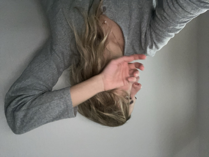 A woman with blonde hair wears a gray long-sleeved shirt. Her face is partially obscured by her hand. The backdrop is a simple, light-colored wall. The image is inverted, creating an unusual perspective. Her eyes are visible, and a necklace is present around her neck.