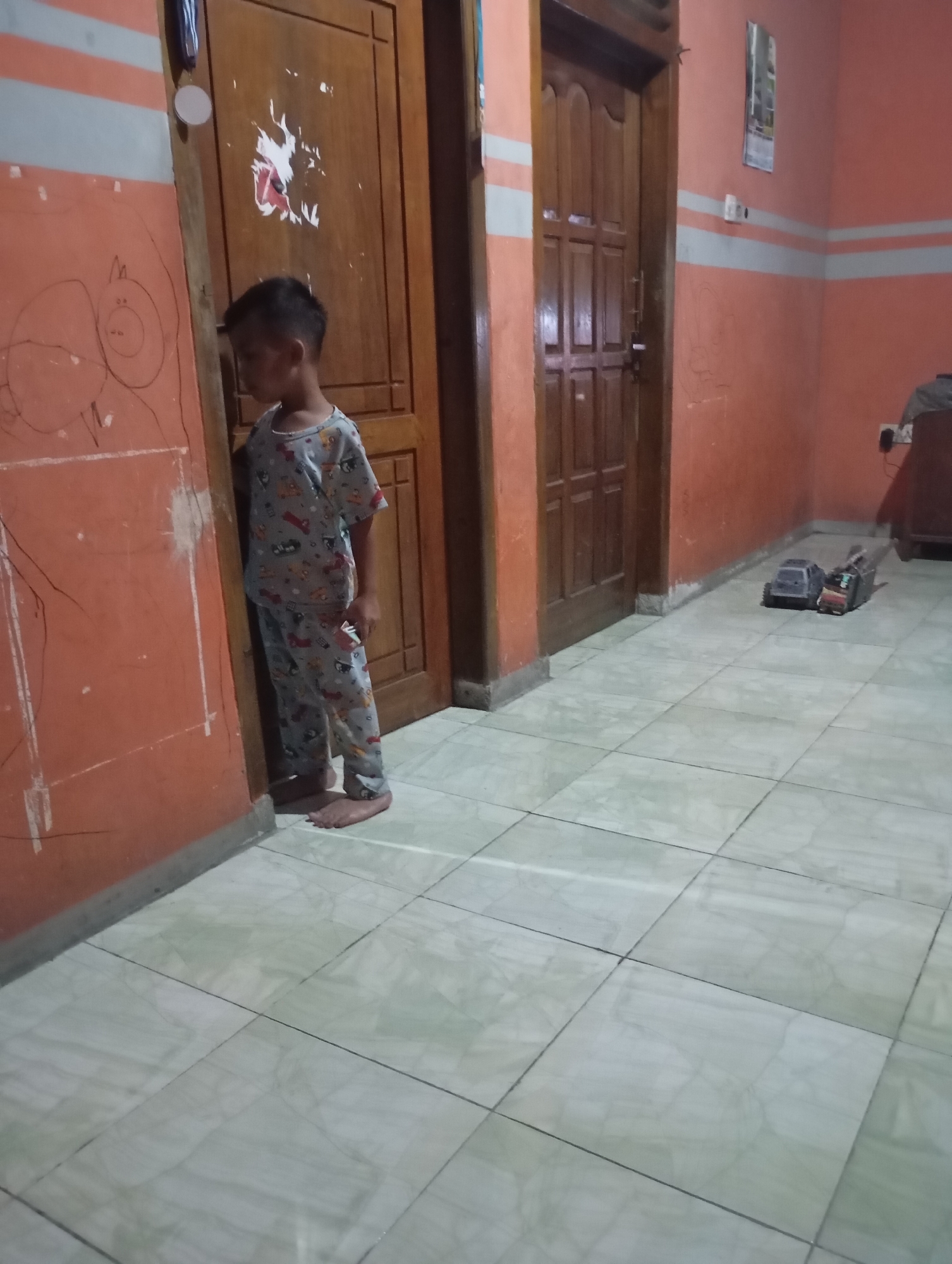 The image depicts an indoor scene, likely a hallway or room, in Ketepung, Indonesia. A young boy, appearing to be of Indonesian descent, stands barefoot and facing a wall. He is wearing pajamas adorned with a pattern of cartoon characters. The wall the boy is facing has child-like drawings on it, suggesting it is a space frequented by children. To the boy's right are two wooden doors, one slightly ajar, revealing a glimpse of an interior space. The floor is tiled in a light-colored pattern. In the background, on the right side of the image, are two toy trucks placed on the tiled floor. The walls are painted an orange hue with horizontal light gray stripes. A calendar is visible on the wall further down the hallway, and an electrical outlet is present near the floor. The lighting suggests it is daytime, but the interior is somewhat dimly lit.