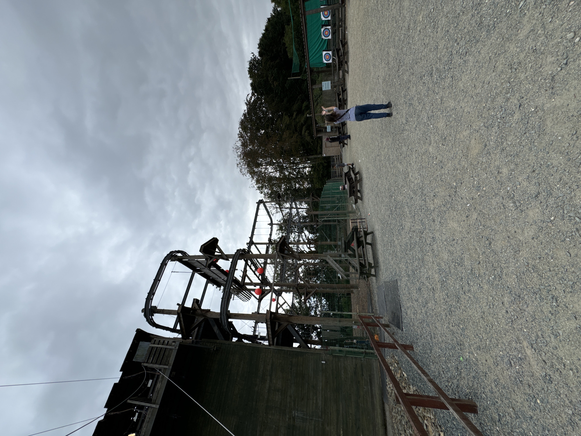 The image depicts an outdoor adventure park on a cloudy day. In the foreground, a gravel ground is visible with a wooden fence on the left. To the left and center, a large wooden structure with a rope course and zipline infrastructure is prominent. Red spherical objects are attached to the rope course. In the background, several picnic tables are scattered. To the right, an archery range is set up with targets. A woman in a striped shirt and jeans is standing in the middle ground, facing away from the camera with her arms raised. Other individuals are present in the background near the archery range and picnic tables. The location is identified as Courtown, Ireland.