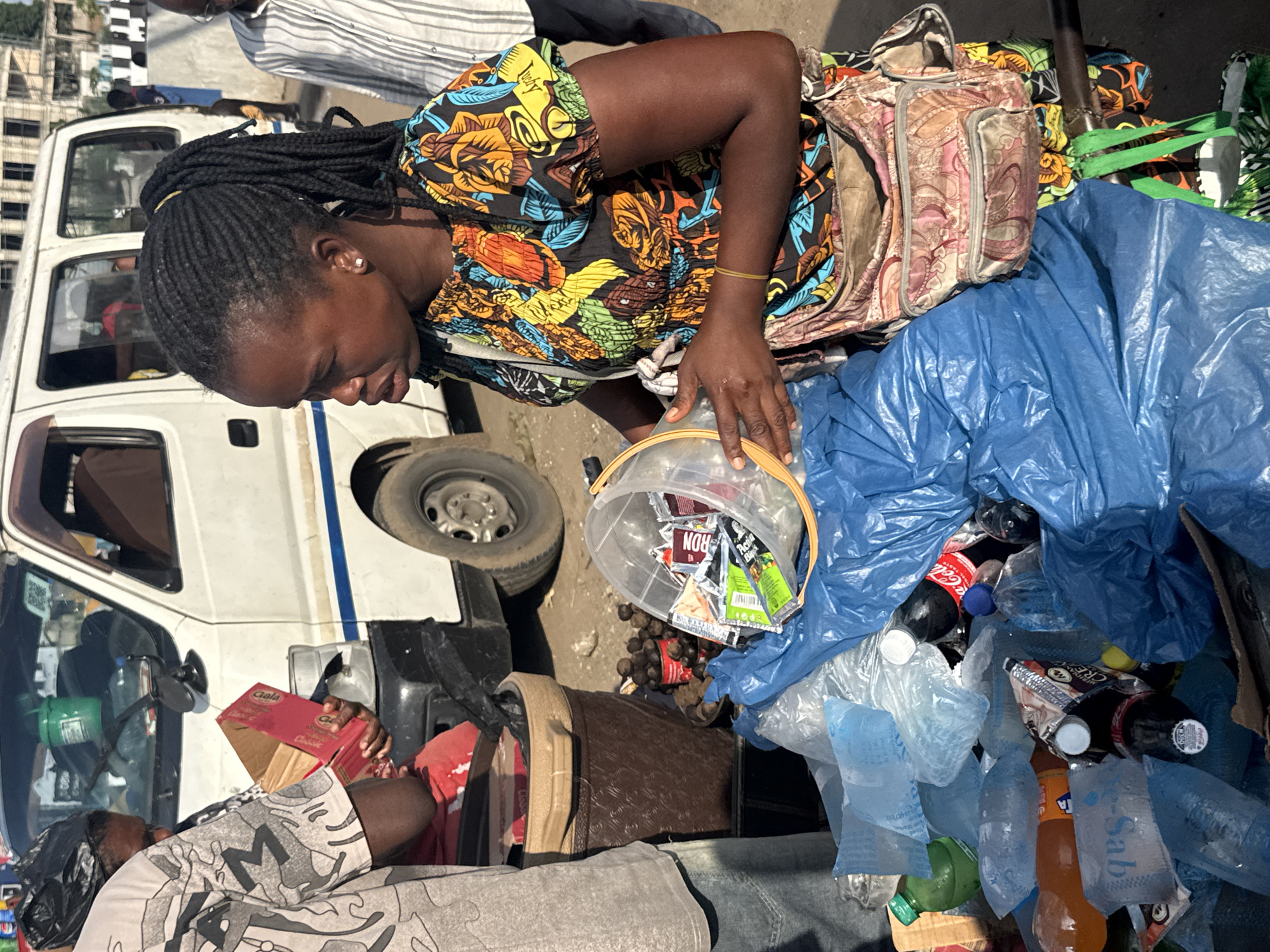 In Owerri, Nigeria, a woman with dark skin and braided hair tied in a ponytail stands beside a white van. She wears a vibrant, colorful dress and a patterned bag slung across her body. In her right hand, she holds a clear plastic bucket filled with various small packages and items. In front of her, a large blue plastic bag spills over with plastic bottles, some empty, some filled with liquid, including a Coca-Cola bottle. To her left, another person, partially visible, holds a red cardboard box. The ground appears dusty and uneven, suggesting a street or marketplace setting. The background shows the white van, its tires, and part of the vehicle's side, along with other people and structures further in the distance.