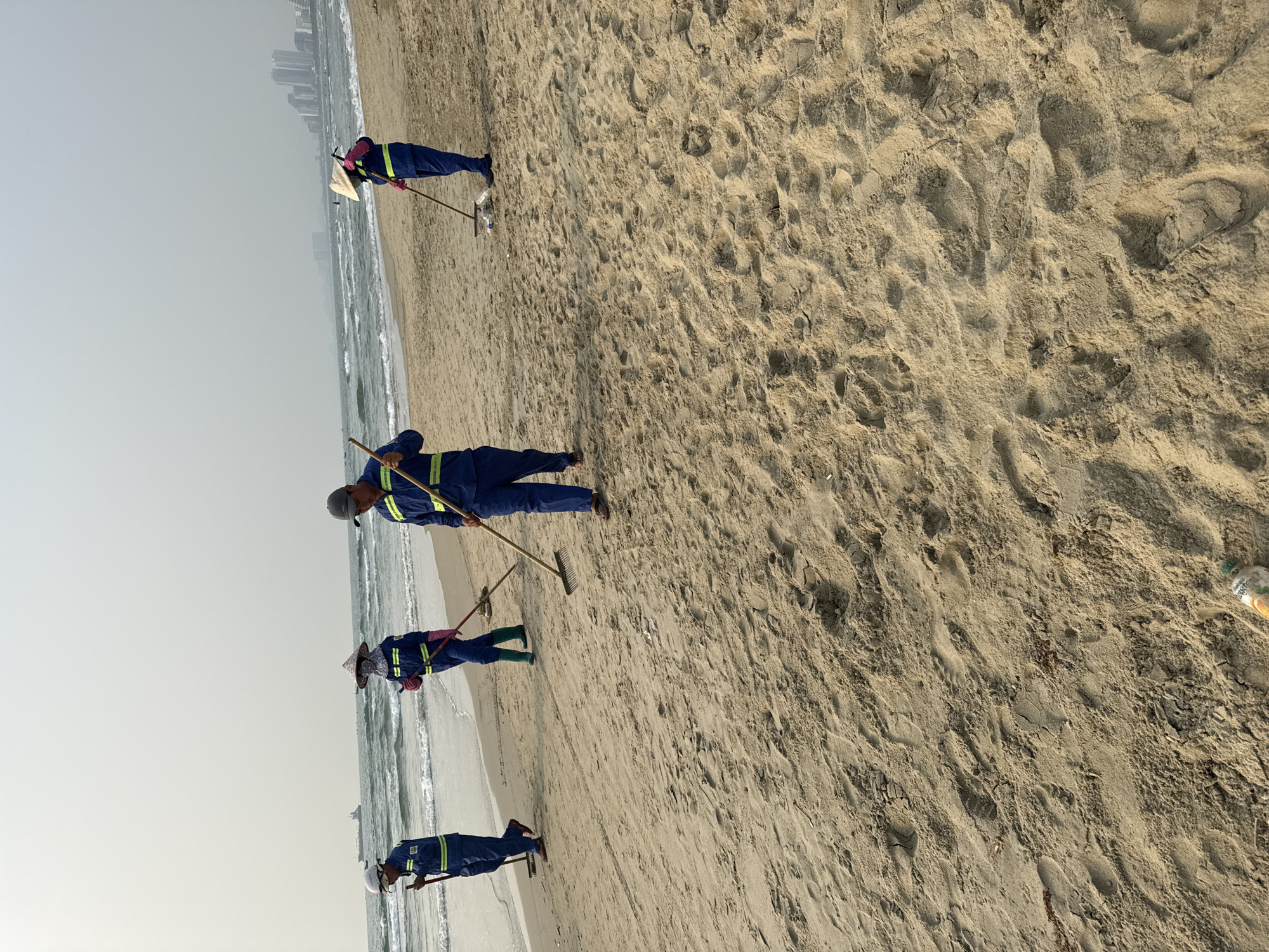 The image depicts a beach scene in Đà Nẵng, Vietnam. Four people wearing blue uniforms with reflective stripes are cleaning the beach. They are using rakes to collect debris. The beach has tan sand, and the ocean is visible in the background, with small waves. A city skyline is faintly visible in the distance. The sky is hazy.