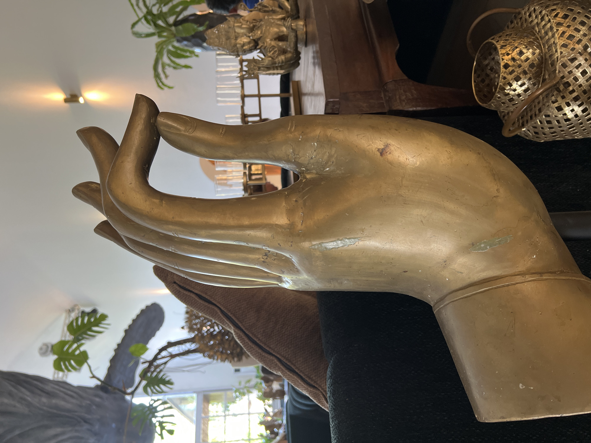 The image displays a large, golden-colored statue of a hand in a pose associated with blessing or peace, a detail likely connected to the cultural significance in Petulu, Indonesia. The hand rests upon a dark-colored surface, which could be a cushion or part of a piece of furniture, with hints of a brown fabric nearby. In the background, a blurred interior setting is visible, featuring plants, a window suggesting natural light, and other decorative objects like a metallic lantern and a smaller statue. The overall scene suggests an indoor setting, potentially a place of worship or a personal shrine, where the statue serves as a central element.