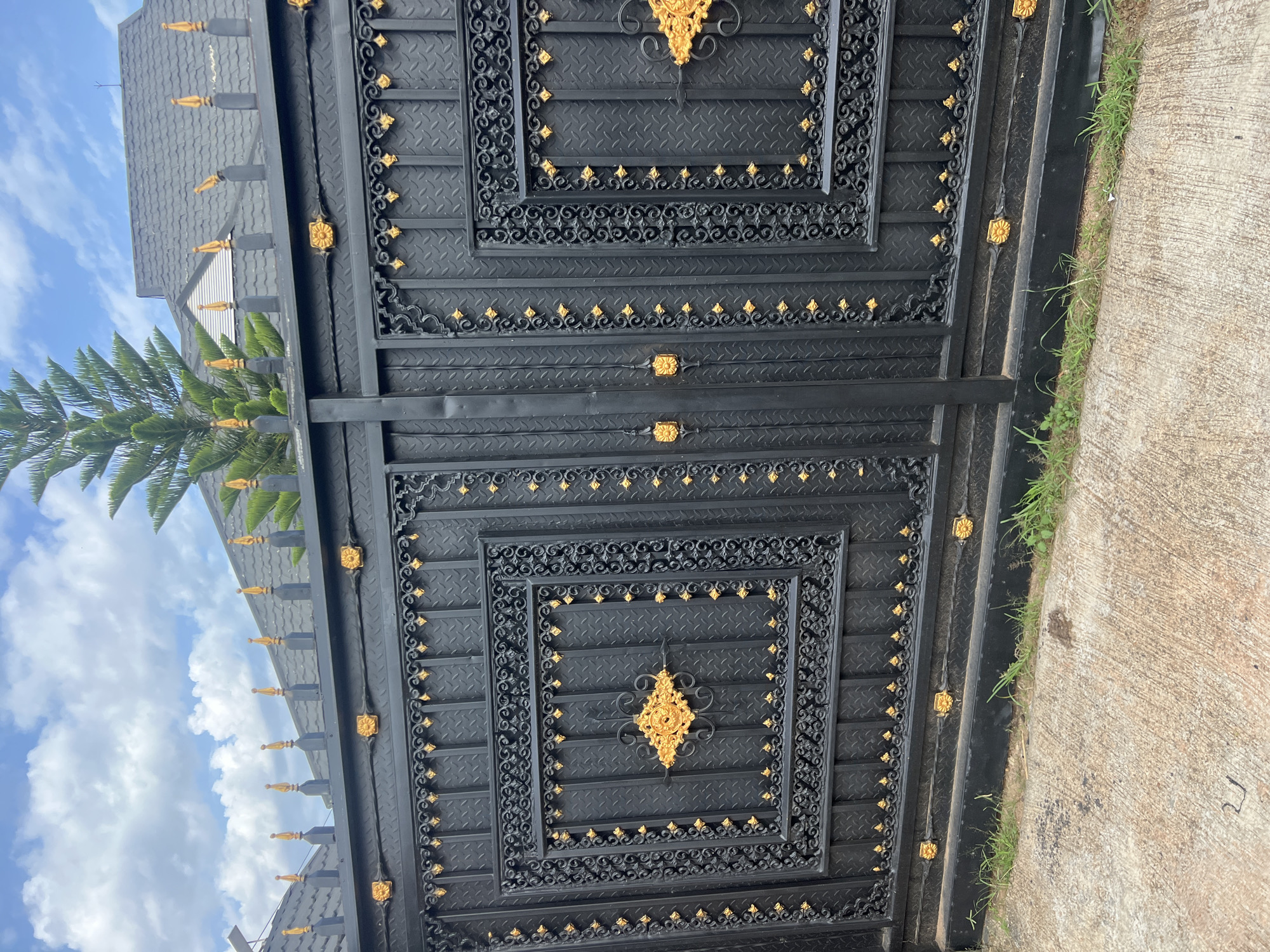 The image shows a large, ornate black metal gate with gold accents. The gate is adorned with intricate scrollwork and diamond-patterned panels. There are two main sections to the gate, each featuring a central square panel with more elaborate gold ornamentation. The gate is topped with decorative finials. In the background, a grey tiled roof of a building is visible, with a tall green pine tree to the right. The sky is blue with scattered white clouds. The gate is situated in front of a concrete driveway with a strip of green grass along the bottom edge. This scene is likely located in the city of Jos, Nigeria, a region known for its cooler climate and the prevalence of pine trees. The image captures a static view of the gate, with no people or activity depicted.