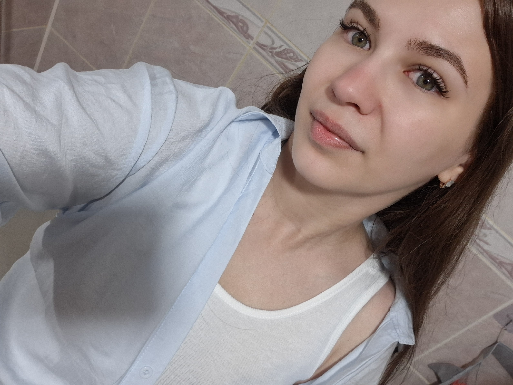 A woman is shown in a selfie-style photograph. She has light skin, green eyes, and brown hair. She is wearing a light blue button-down shirt over a white tank top. There is a patterned tile wall in the background. The photo does not provide enough information to identify a specific location in the city of Chaikovsky, Russia.
