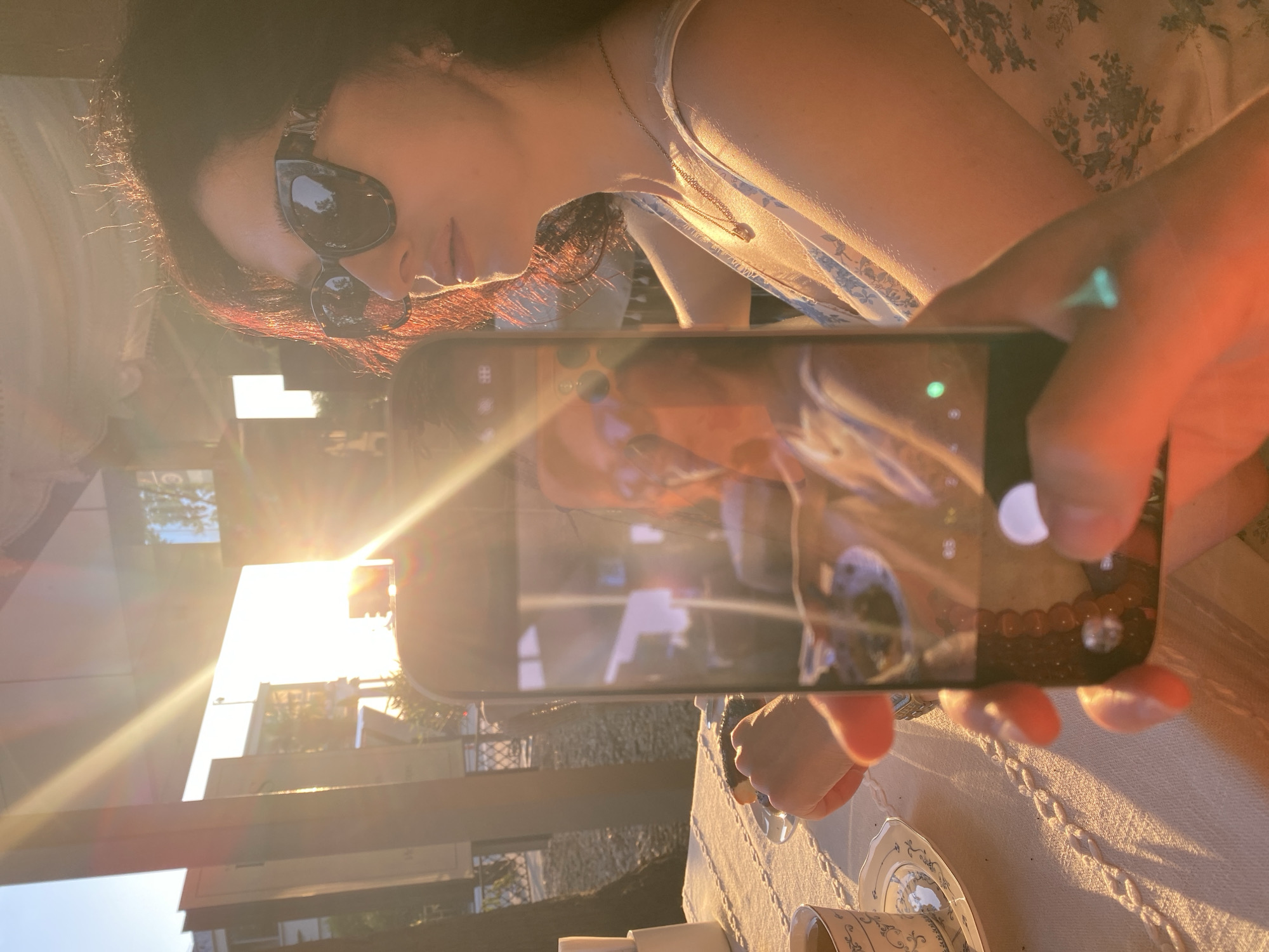 The image captures an outdoor cafe scene in Sochi, Russia, under strong, warm, low-angle sunlight, indicating the golden hour. In the foreground, a person's hands, with manicured fingernails, hold a dark-colored smartphone in portrait orientation, actively utilizing its camera function. The phone's screen displays a camera interface, including a visible shutter button, and reflects parts of the surrounding environment.
The primary subject, positioned to the right of the frame, is a woman with dark, curly hair and dark, patterned cat-eye sunglasses. She wears a light-colored, sleeveless dress with a blue floral pattern and a delicate gold-toned necklace. Her head is turned to her right (viewer's left), and she is illuminated from behind and the side by the intense sunlight, which creates significant lens flare and backlights her hair. Her right arm is visible, resting on a table covered with a white, textured tablecloth featuring a chain-link pattern.
On the table, a white teacup with a matching saucer, both adorned with intricate blue and gold designs, is placed to the left. A dark, cylindrical pastry or dessert rests on a small white plate near the woman's right hand. The background includes trees, various architectural structures, and a light sky, consistent with an urban outdoor setting or patio.