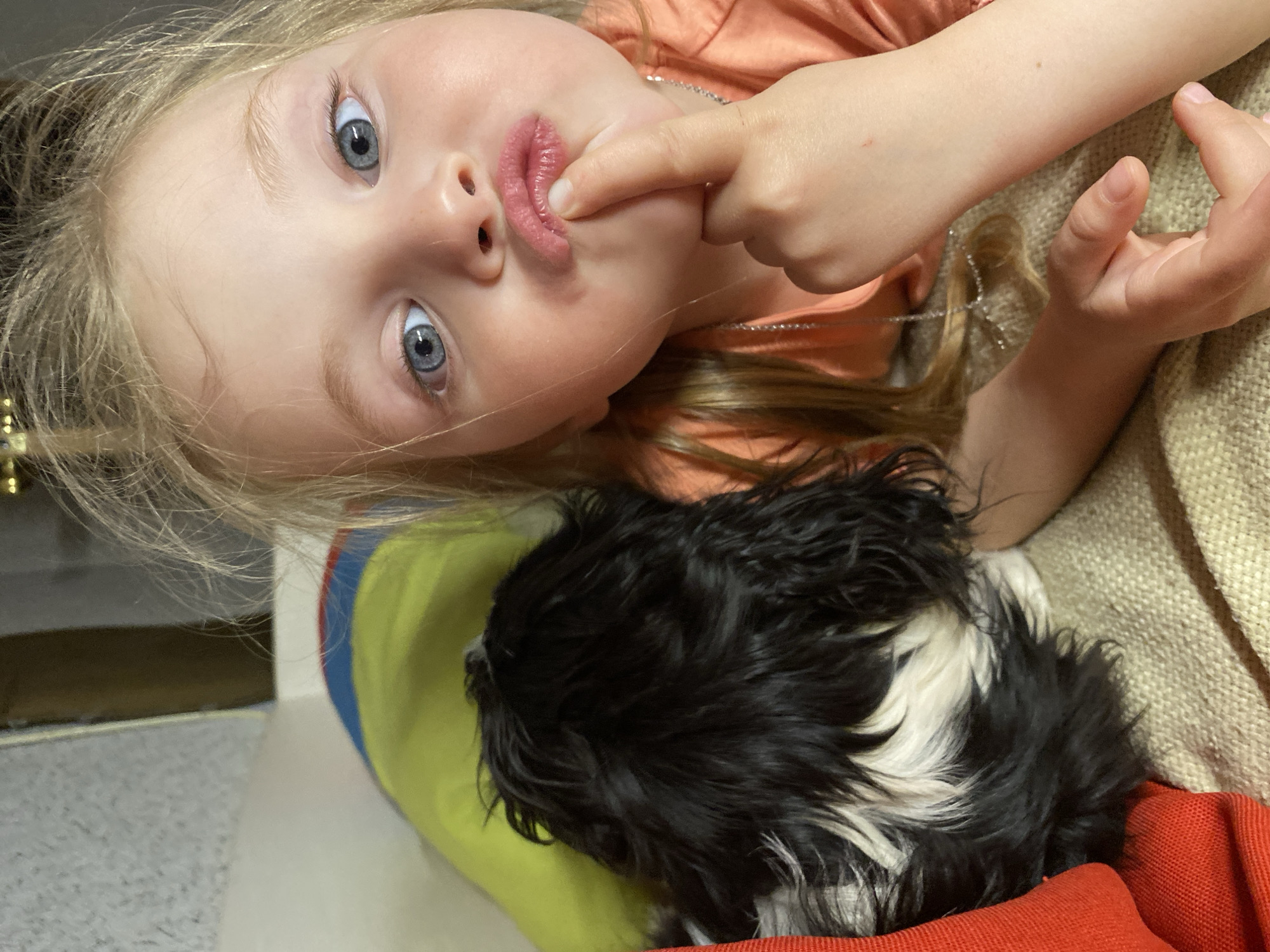A young child with blonde hair and blue eyes is shown from an inverted perspective, lying on a couch with a black and white dog. The child is wearing an orange shirt and a silver necklace. The child has one finger touching their lips. The dog is lying on a colorful cushion (red, yellow, and blue) and a beige throw is partially visible. The location is not evident in the image; there is no indication that the location is in Votkinsk, Russia.