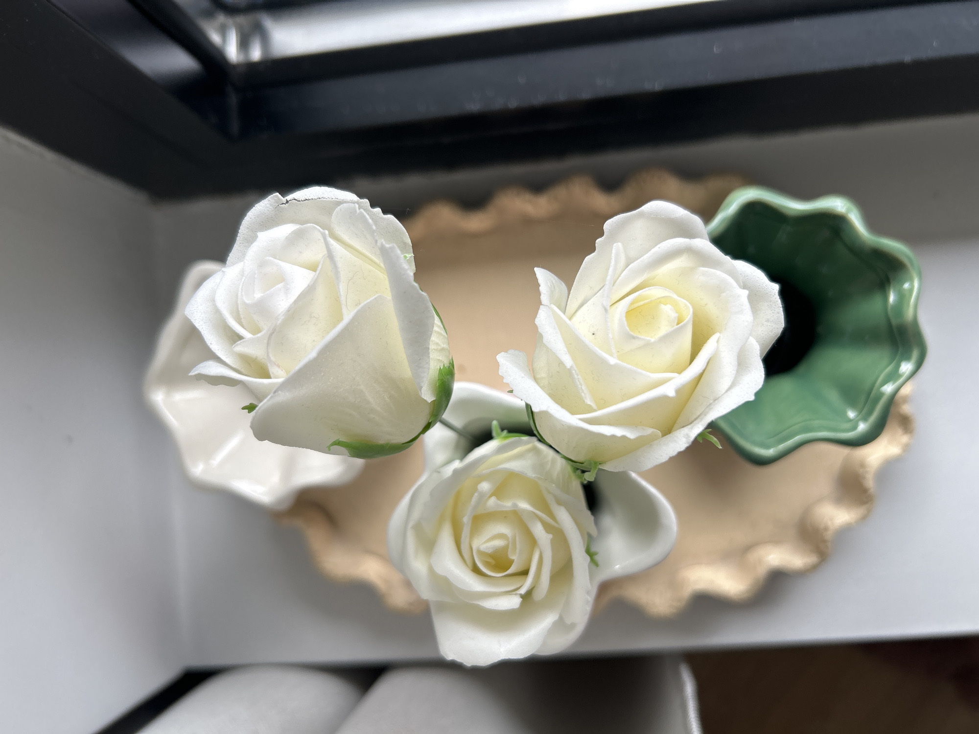 The image shows three artificial white roses arranged in small, decorative dishes on a beige scalloped tray. One dish is white, and the other is green. The background includes a black frame, possibly a window frame or a shelf. The scene appears to be indoors and there is no evidence indicating this image was taken in Sofia, Bulgaria.