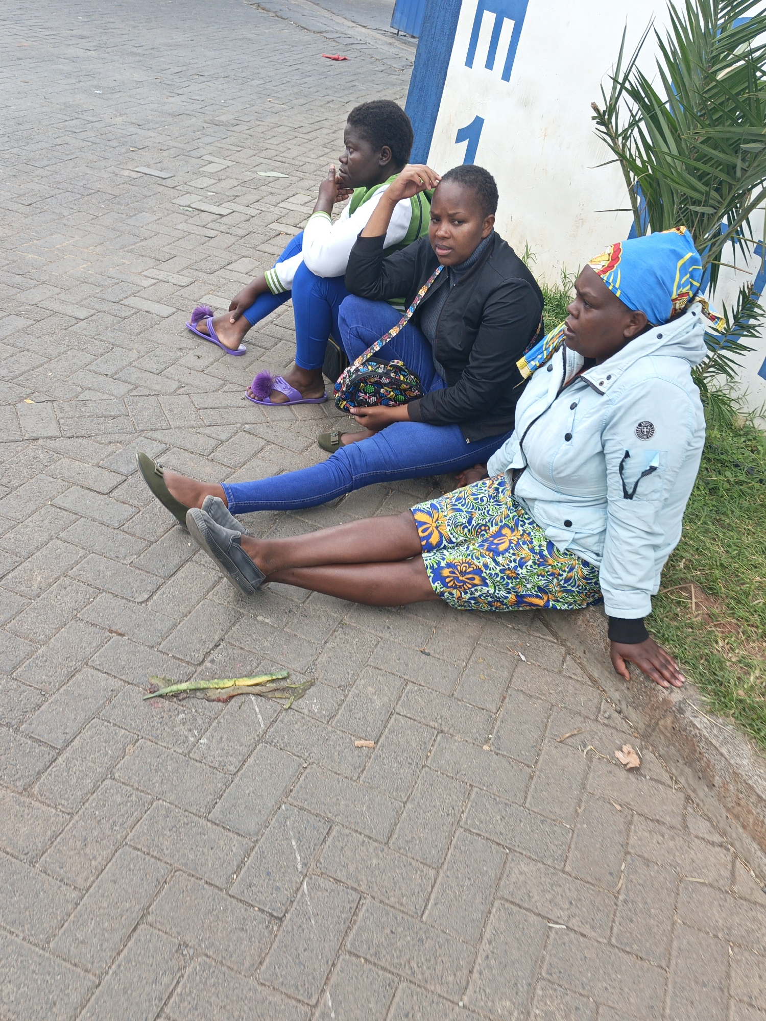 Three Black women are sitting on a brick-paved sidewalk near a wall. The woman furthest to the left wears a white and green jacket, blue jeans, and purple sandals. The woman in the middle wears a black jacket, blue jeans, and carries a patterned handbag. The woman to the right is wearing a light blue jacket, a blue floral skirt, and a blue headscarf. All three women are looking away from the camera. A plant is visible behind them. The location is the city of Nairobi, Kenya.