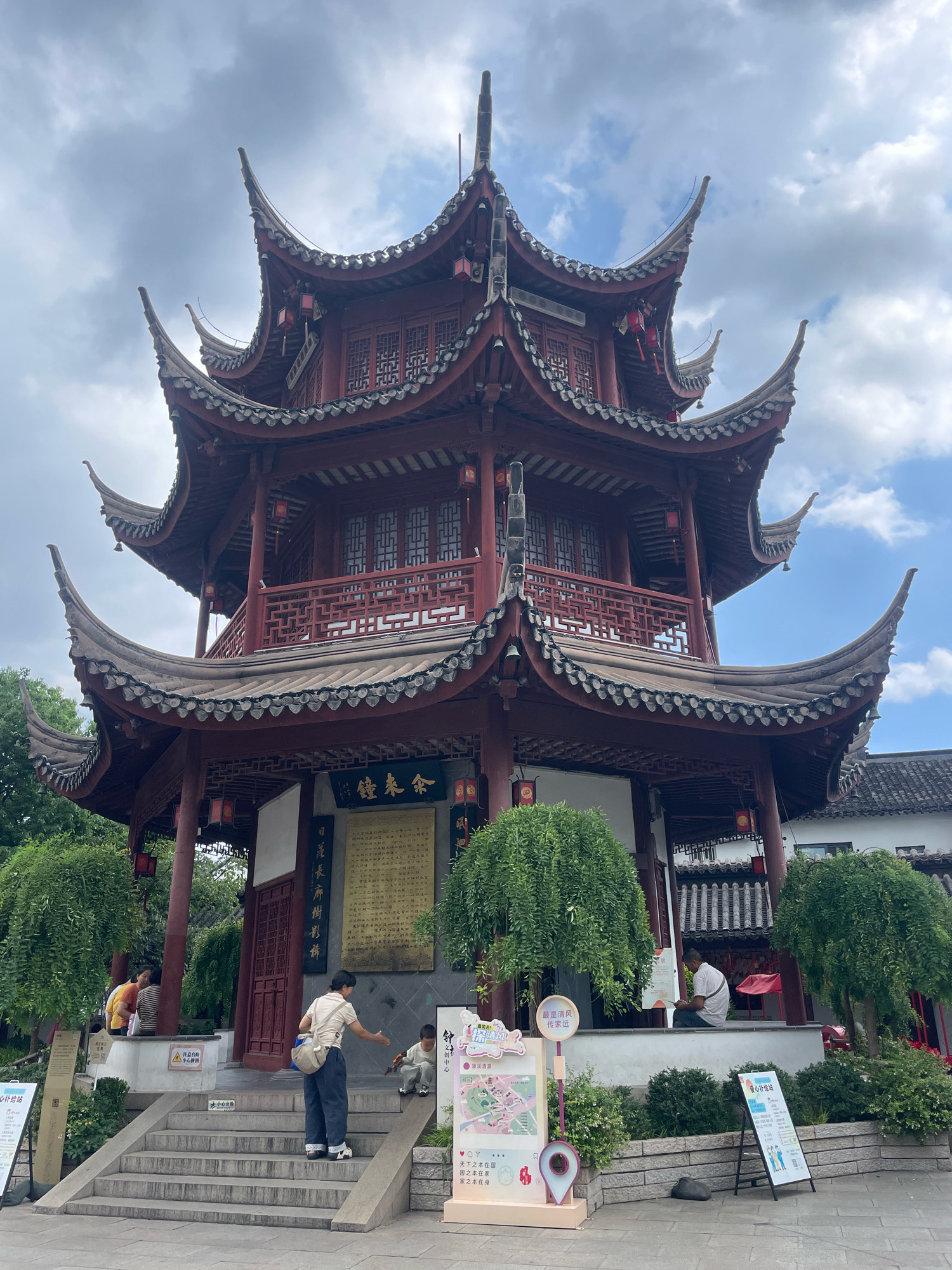 An outdoor, medium-shot, eye-level, frontal view captures a multi-tiered, traditional Chinese pavilion, painted crimson red with ornate grey tile roofs, situated in Minhang District, China. The pavilion's prominent features include intricate latticework on balconies and windows, as well as decorative red lanterns hanging from its eaves. Beneath the pavilion, a grey stone plaque inscribed with Chinese characters is mounted on a dark wall.
In the foreground, a concrete staircase leads up to the pavilion's entrance. On the stairs, a woman wearing a beige t-shirt, dark pants, and a shoulder bag gestures with her right hand towards a young boy who is kneeling and reaching for something on the ground. The boy is dressed in grey. To the left of the staircase, a woman in a yellow shirt and a person in a striped shirt are seated on a ledge.
To the right of the staircase, a tall, colorful informational display with a map is positioned. Further to the right, a man in a white shirt is seated on a ledge, looking down at his mobile phone. Two decorative, rounded trees flank the pavilion's lower level. Lush green bushes are visible in the background. The sky above is partly cloudy, with a mix of blue sky and grey clouds.