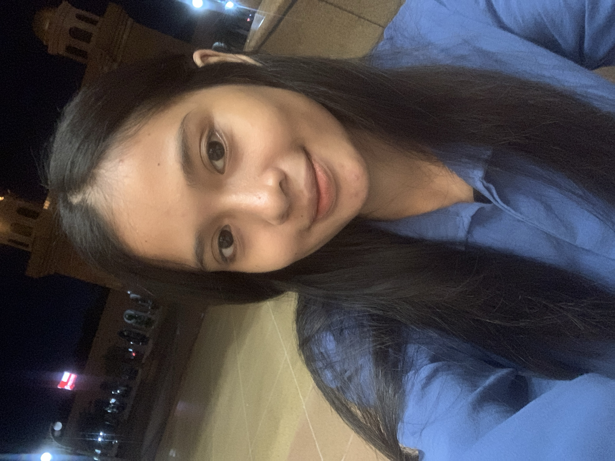 The image is a selfie-style portrait of a young woman taken at night, rotated clockwise. The primary subject is a young woman with long, dark hair and dark eyes, looking directly at the camera with a subtle, pleasant smile. She is wearing a blue long-sleeved top or shirt.
The background is dark, indicating nighttime, with various artificial lights visible in the distance. To the upper left, a faintly illuminated building with distinct architectural features, possibly a tower or a prominent public structure, is visible against the dark sky. Below the woman, a paved ground surface is visible. Additional crops reveal close-ups of her face, hair, and the blue garment, as well as more details of the dark, urban background with scattered bright lights and what appears to be a red and white sign or object in the distance. The scene suggests an outdoor setting, likely a public area or street in Samarinda, Indonesia. No discernible text is visible.