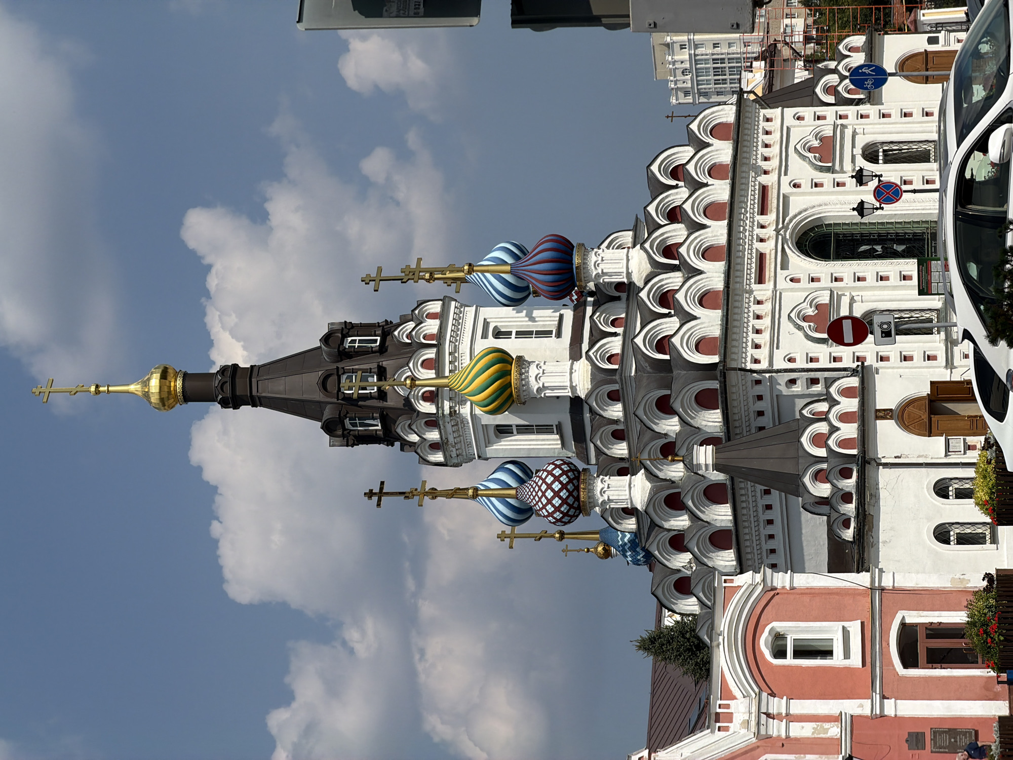 The image shows the Church of the Assumption of the Blessed Virgin Mary in Saratov, Russia, under a cloudy sky. The church is a vibrant example of Russian Orthodox architecture, featuring multiple onion domes of various colors and patterns, including blue and white stripes, green and yellow stripes, and red and white checkered patterns. The main structure is white with red accents and decorative elements. A golden cross tops the tallest spire. To the left, a pink building with white trim is partially visible, with a dark-tiled roof and several windows. In the foreground, the rear of a white car is visible. Several traffic signs are present, including a "no entry" sign with a camera symbol, a pedestrian crossing sign, and another circular sign with a red border and a diagonal bar. The environment appears to be an urban setting with a paved area in the foreground and trees visible.