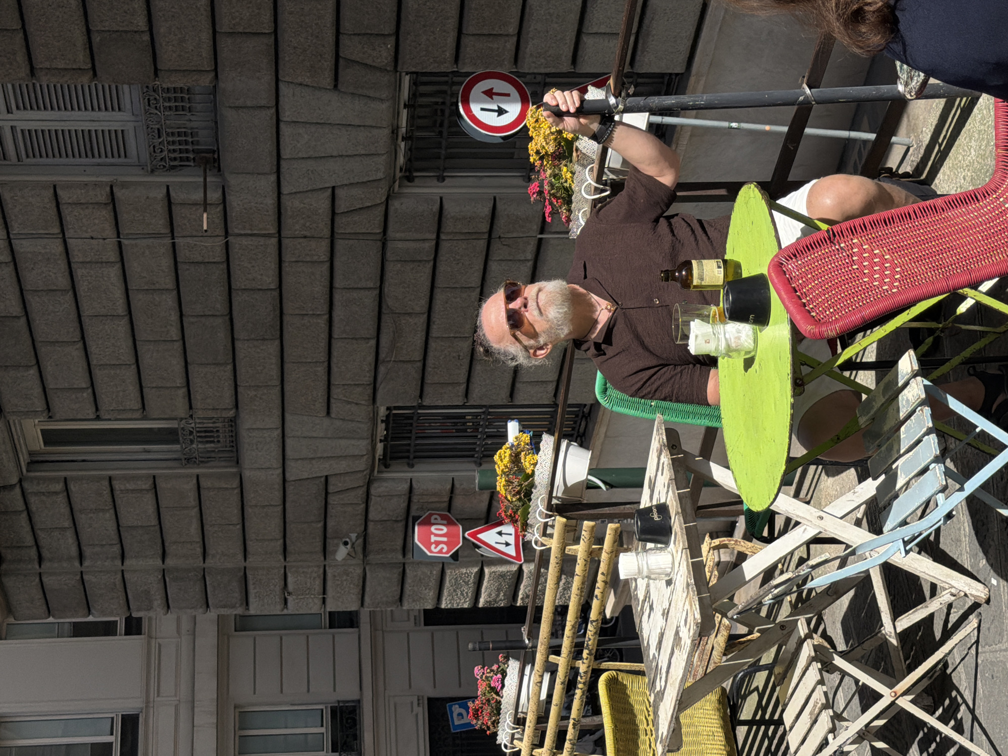 A slightly high-angle shot depicts a man with a white beard seated at an outdoor cafe in Genoa, Italy. He wears a brown short-sleeved shirt and sunglasses and looks up. A green, round table is in front of him, adorned with a brown glass bottle, glass tumblers, and what appears to be crumpled napkins. The man sits on a light green chair. To his right, a red wicker chair is also visible.
The cafe setting includes two tables, one with a light-colored wooden surface and another with a green surface. Street signs indicating traffic directions are visible above the cafe. The cafe is situated against a stone-tiled building facade, and a "Stop" sign can be seen behind the cafe. A security camera is mounted on the building. Other people are present but only partially visible in the frame.
